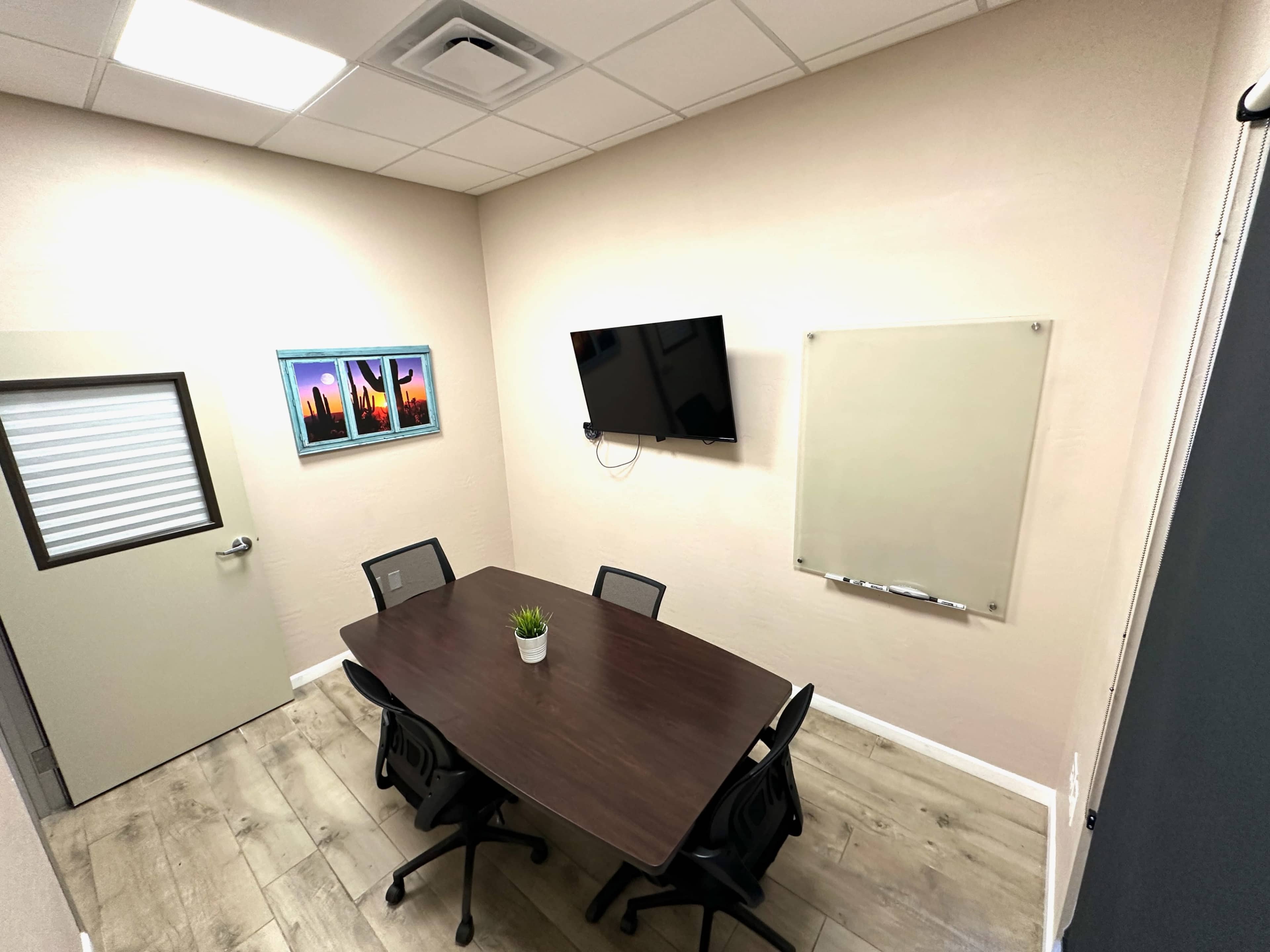 The image shows a small conference room with a rectangular table, four chairs, a wall-mounted TV, a whiteboard, and framed pictures on the wall.