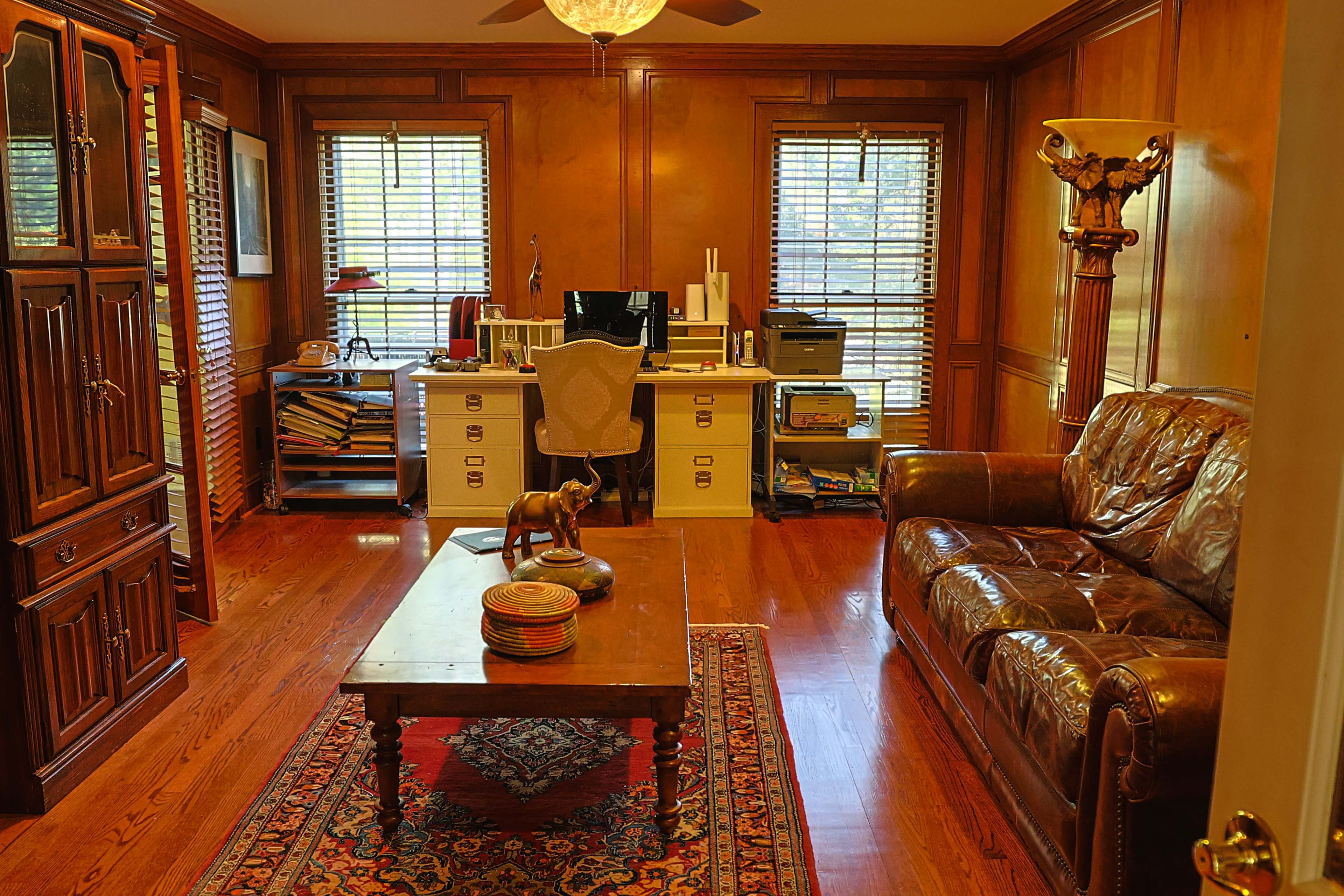 The image shows a wooden-paneled office with a desk, a couch, and a decorative table in the center, featuring a computer and shelves in the background.
