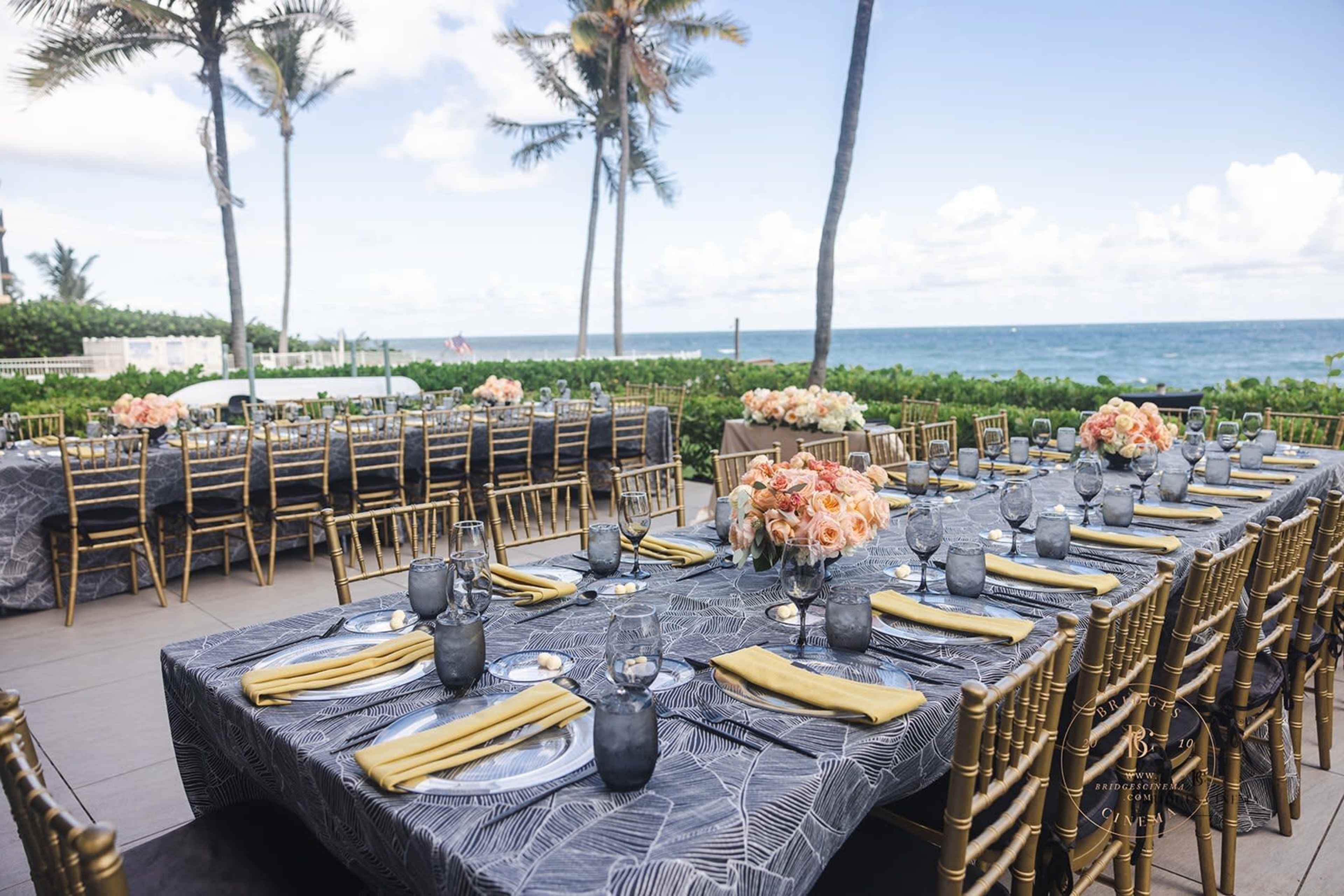 A beautifully arranged outdoor dining setup features multiple tables with gold chairs, set against a backdrop of palm trees and the ocean.