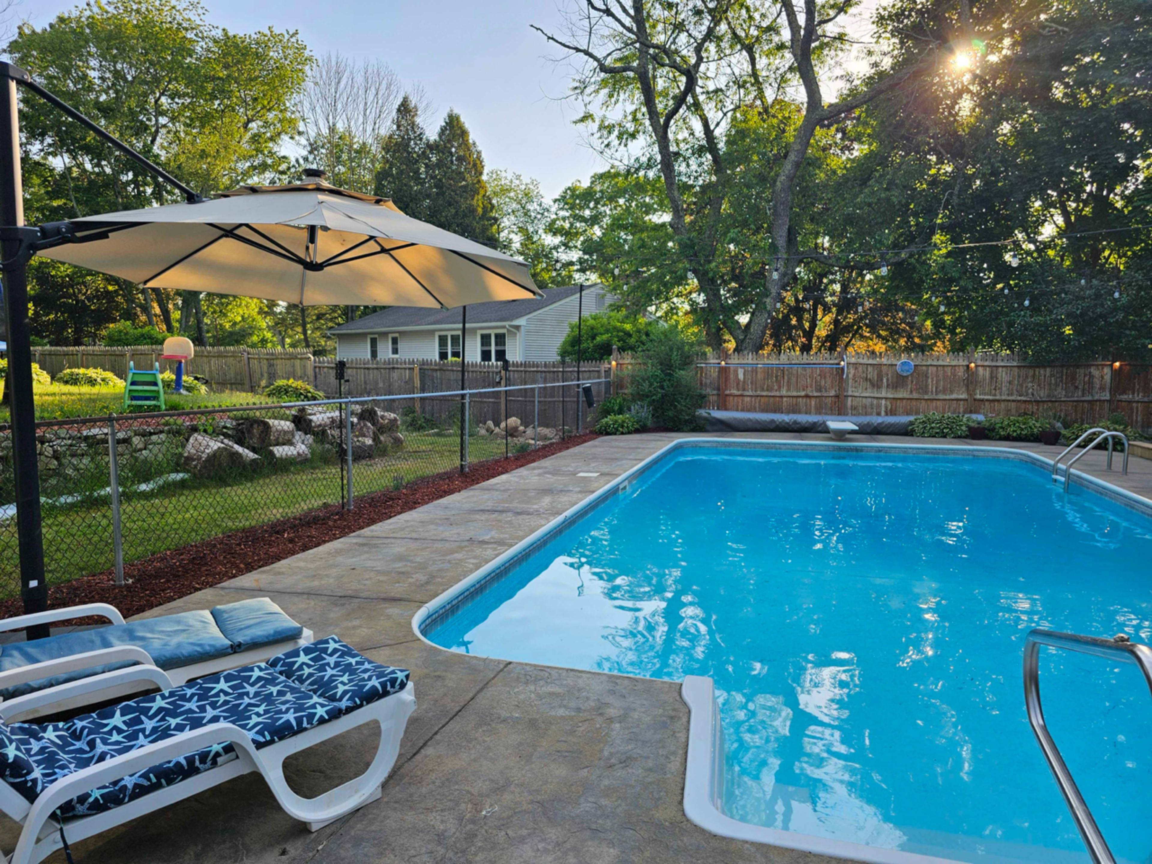 A clear blue pool is surrounded by a patio with lounge chairs and an umbrella, overlooking a fenced backyard with greenery and a playset.