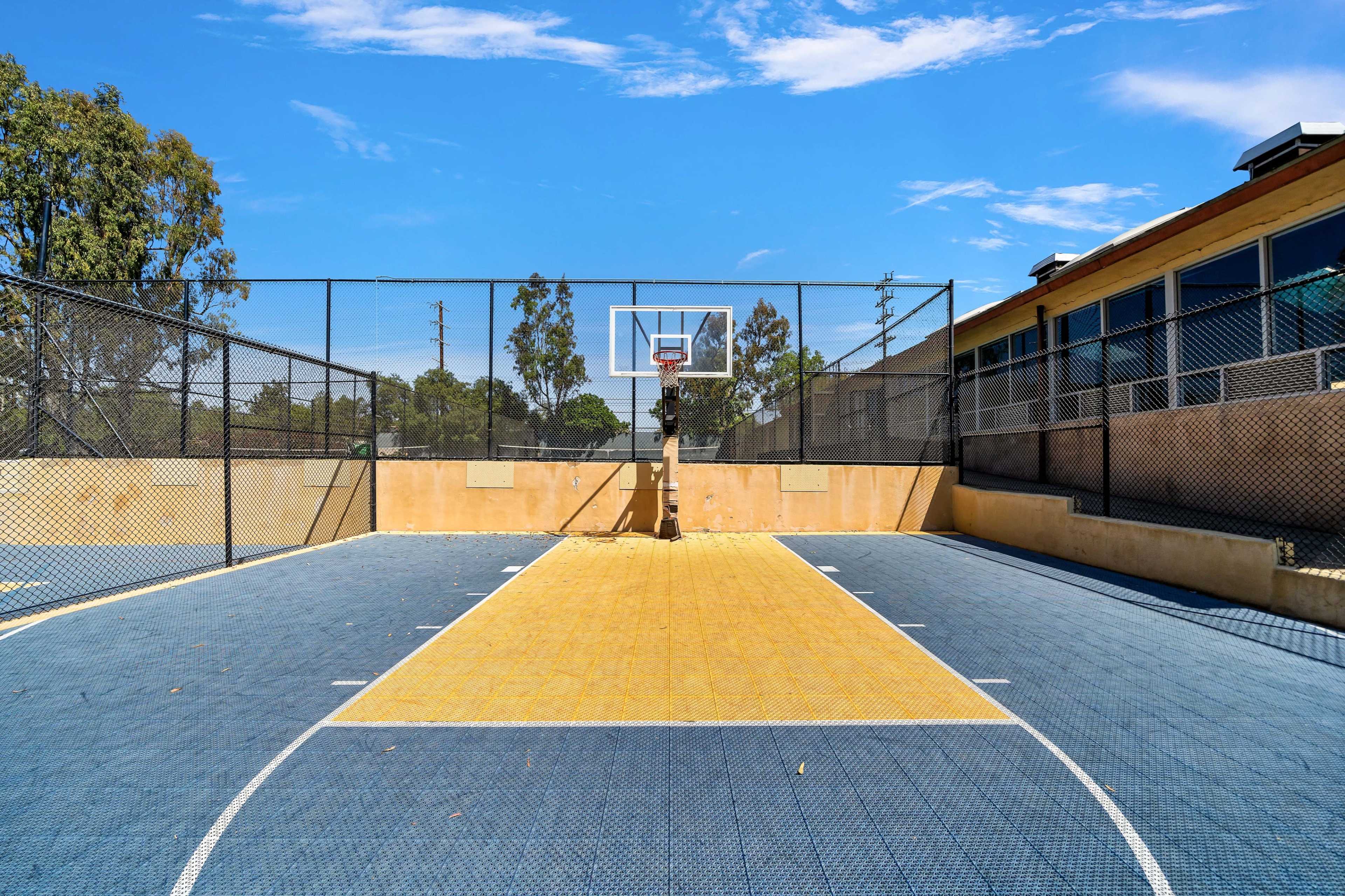 A fenced basketball court with a yellow and blue surface, a hoop at the far end, and trees and a building in the background.