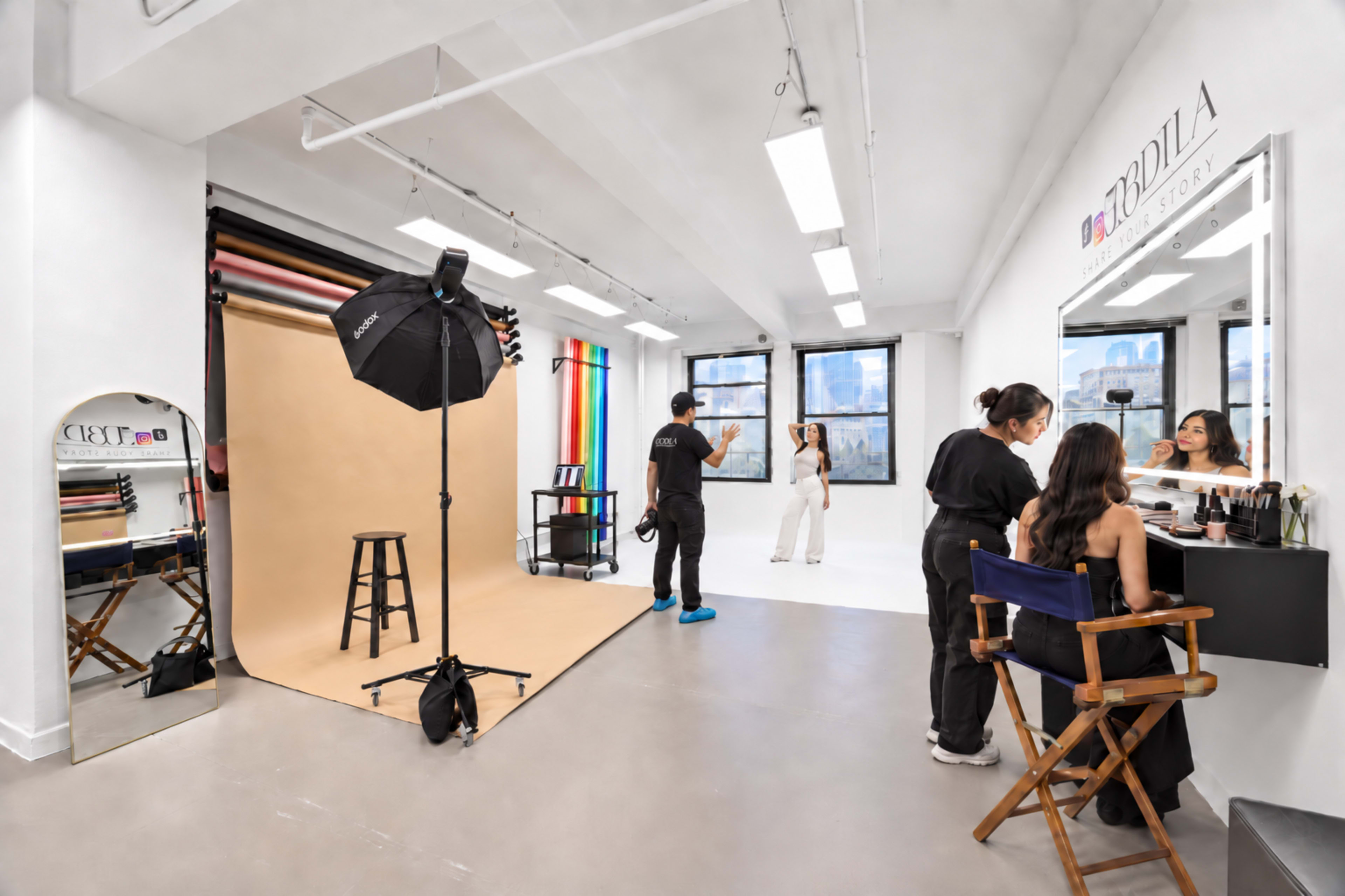 The image shows a photography studio featuring a backdrop setup, lighting equipment, and two women preparing for a photoshoot while another woman sits in front of a makeup mirror.