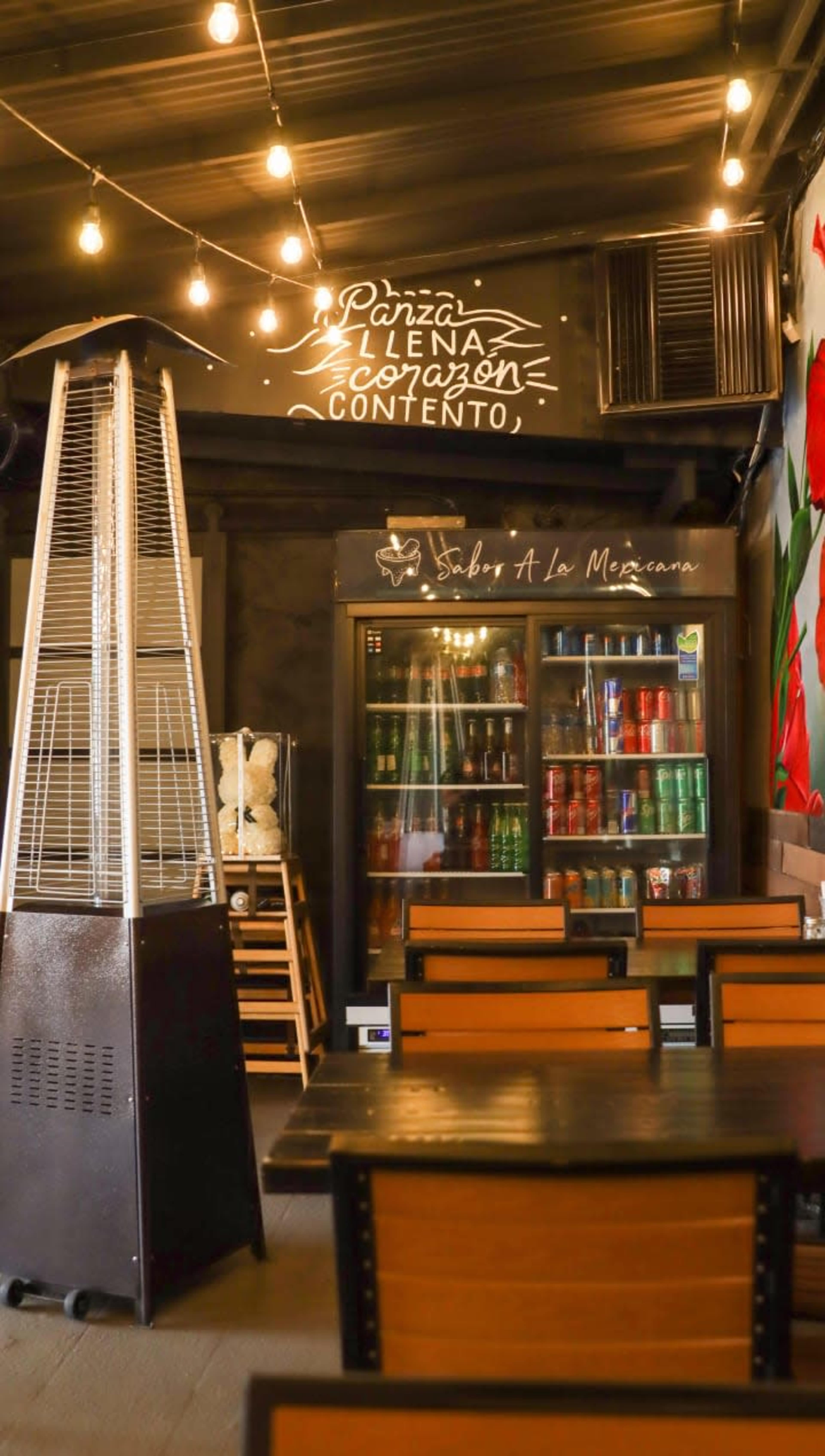 The image shows the interior of a cozy restaurant featuring wooden tables, a glass display refrigerator filled with drinks, and string lights overhead.
