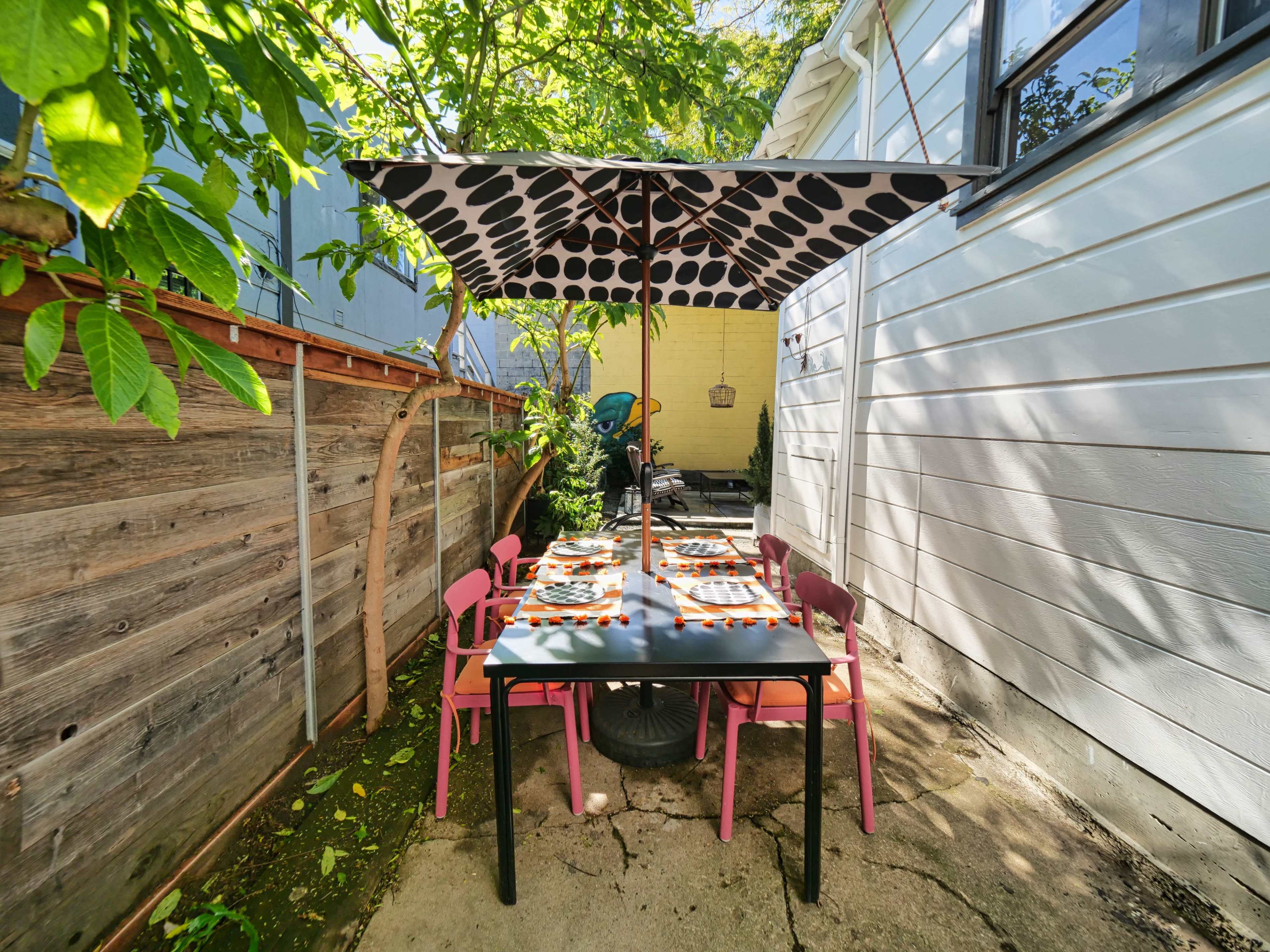 A dining table with colorful plates is set under a polka-dotted umbrella in a shaded outdoor space surrounded by greenery.