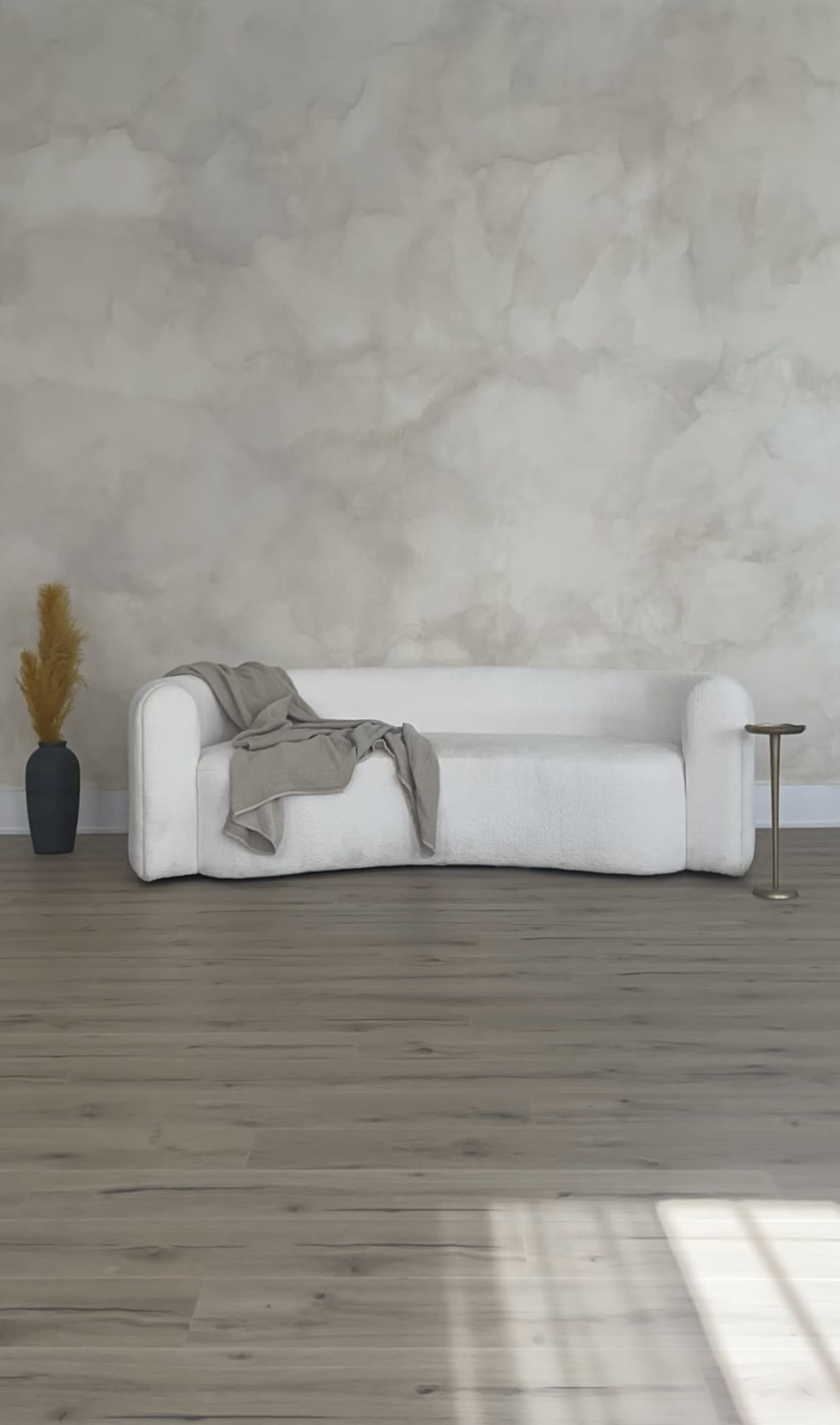 A white, curved sofa with a gray throw blanket is positioned against a textured beige wall, accompanied by a vase and a small side table, on a wooden floor.
