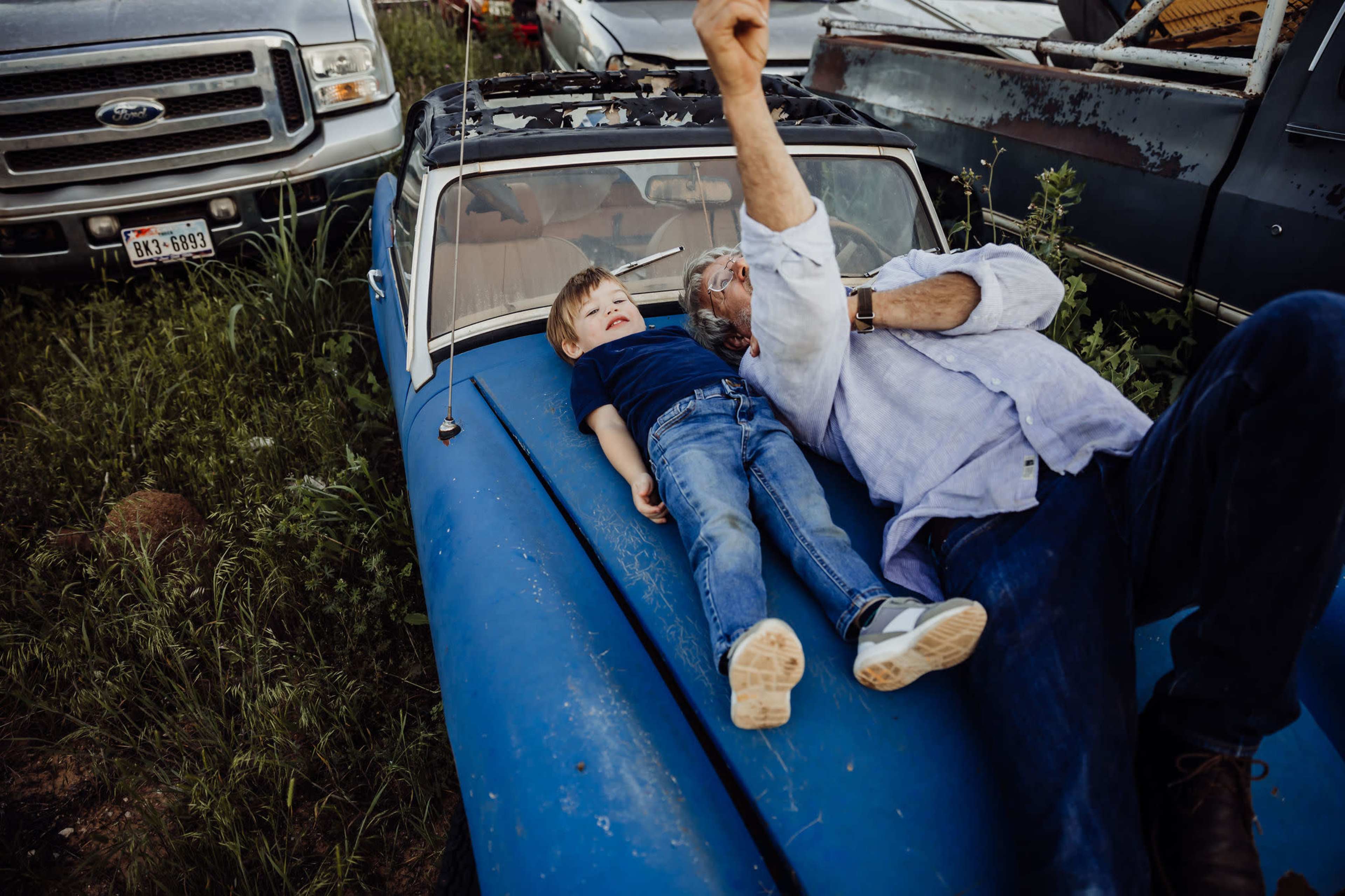 A man and a child lie on the hood of a blue car surrounded by other vehicles in a grassy field.