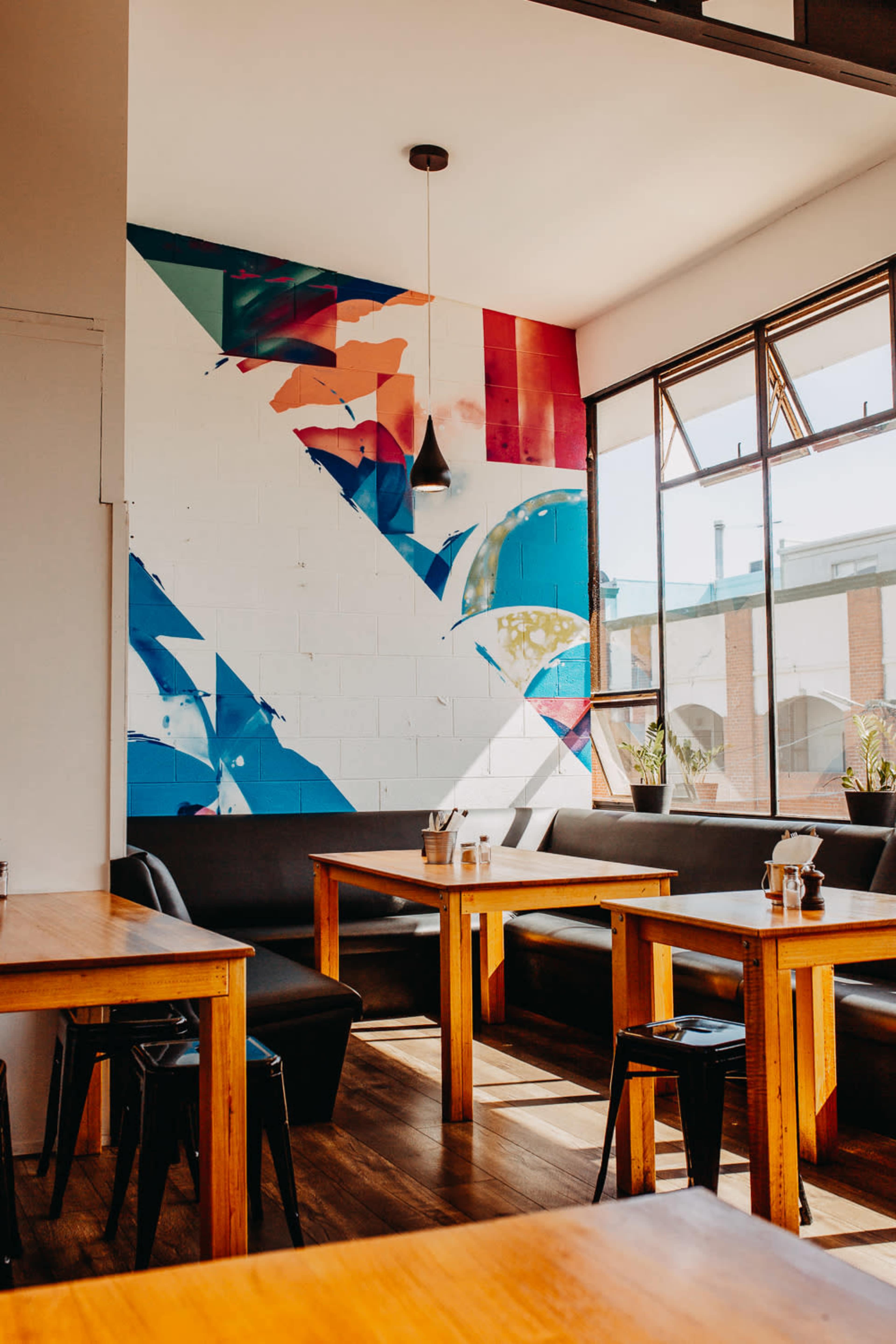 The interior of a restaurant features wooden tables, black seating, and a colorful mural on the wall.