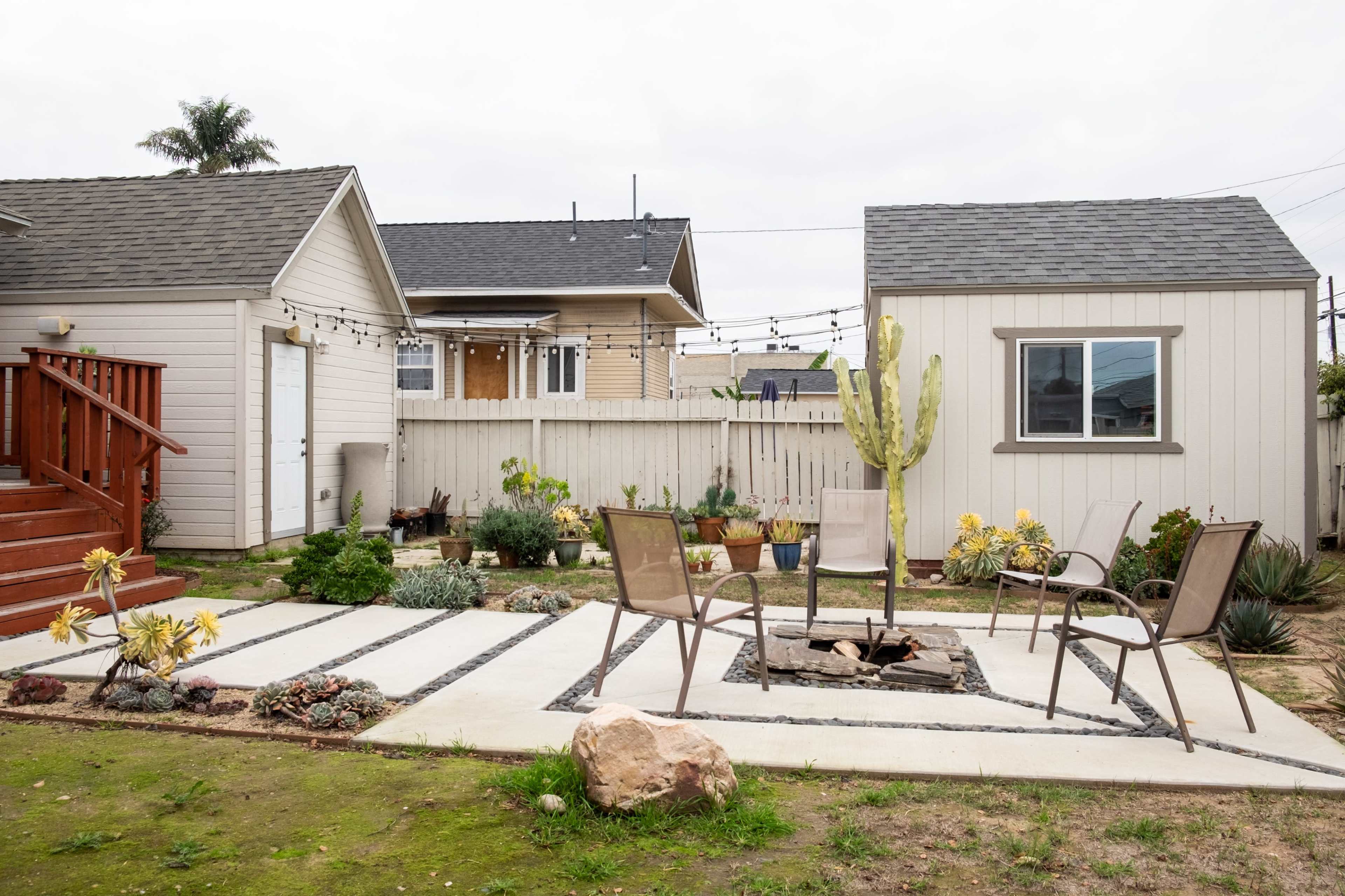 A landscaped backyard featuring a fire pit surrounded by concrete pathways and two chairs, with nearby planters and a cactus.