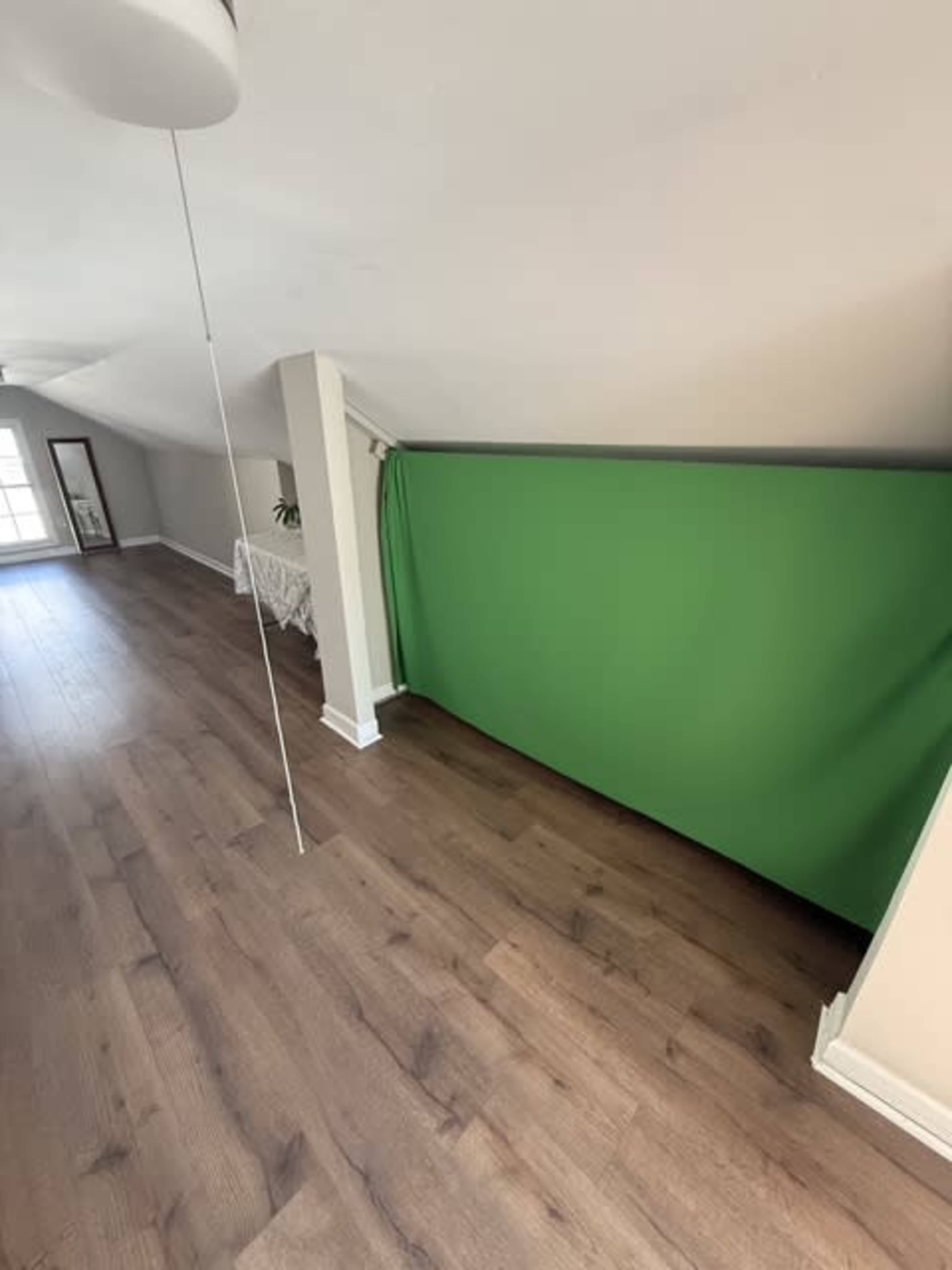 The image shows a spacious room with wooden flooring and a green backdrop against a slanted wall.