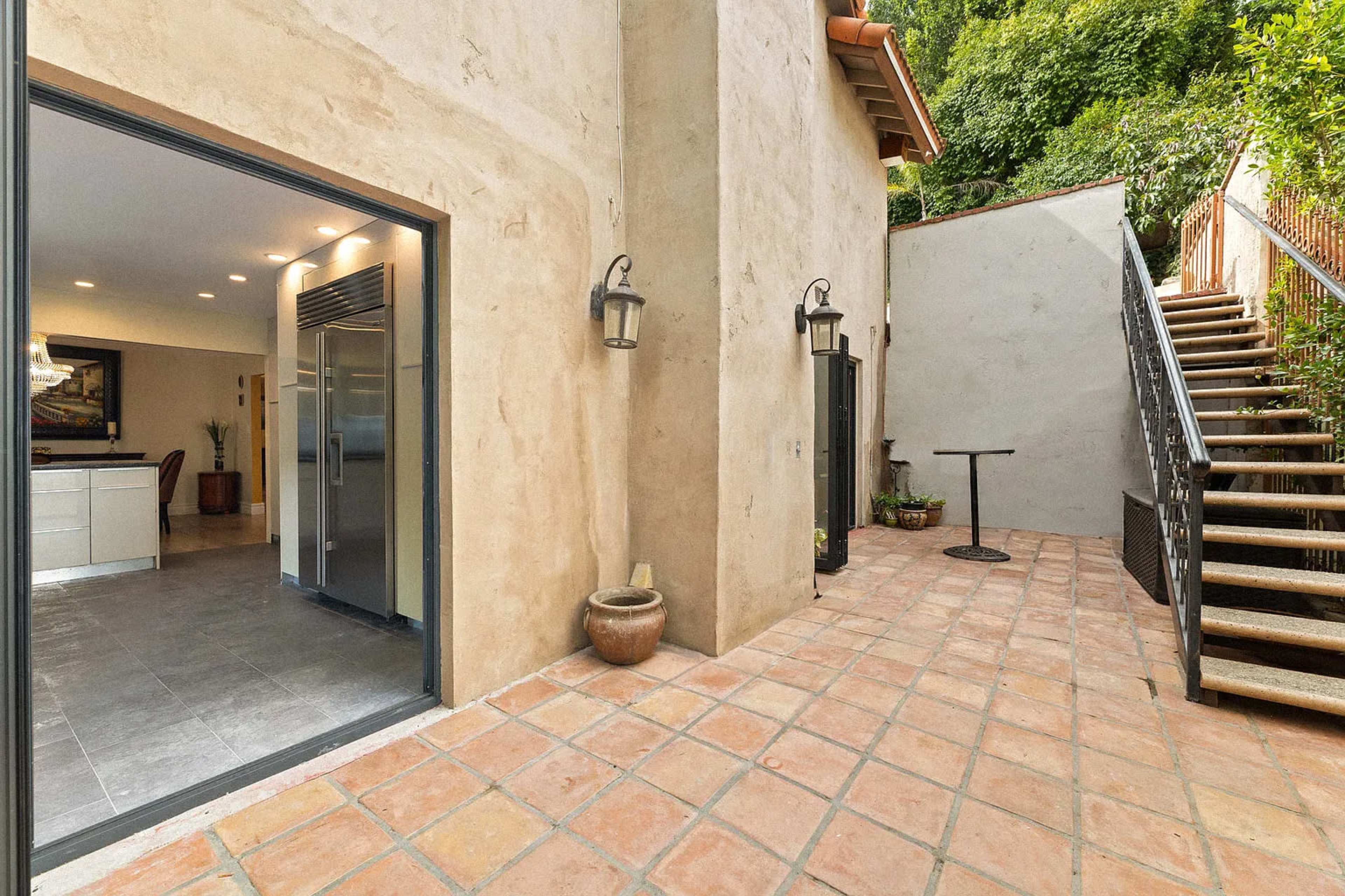 The image shows a patio area with terracotta tiles, a small round table, an outdoor staircase leading to a higher level, and a doorway that opens into a kitchen space with modern appliances.