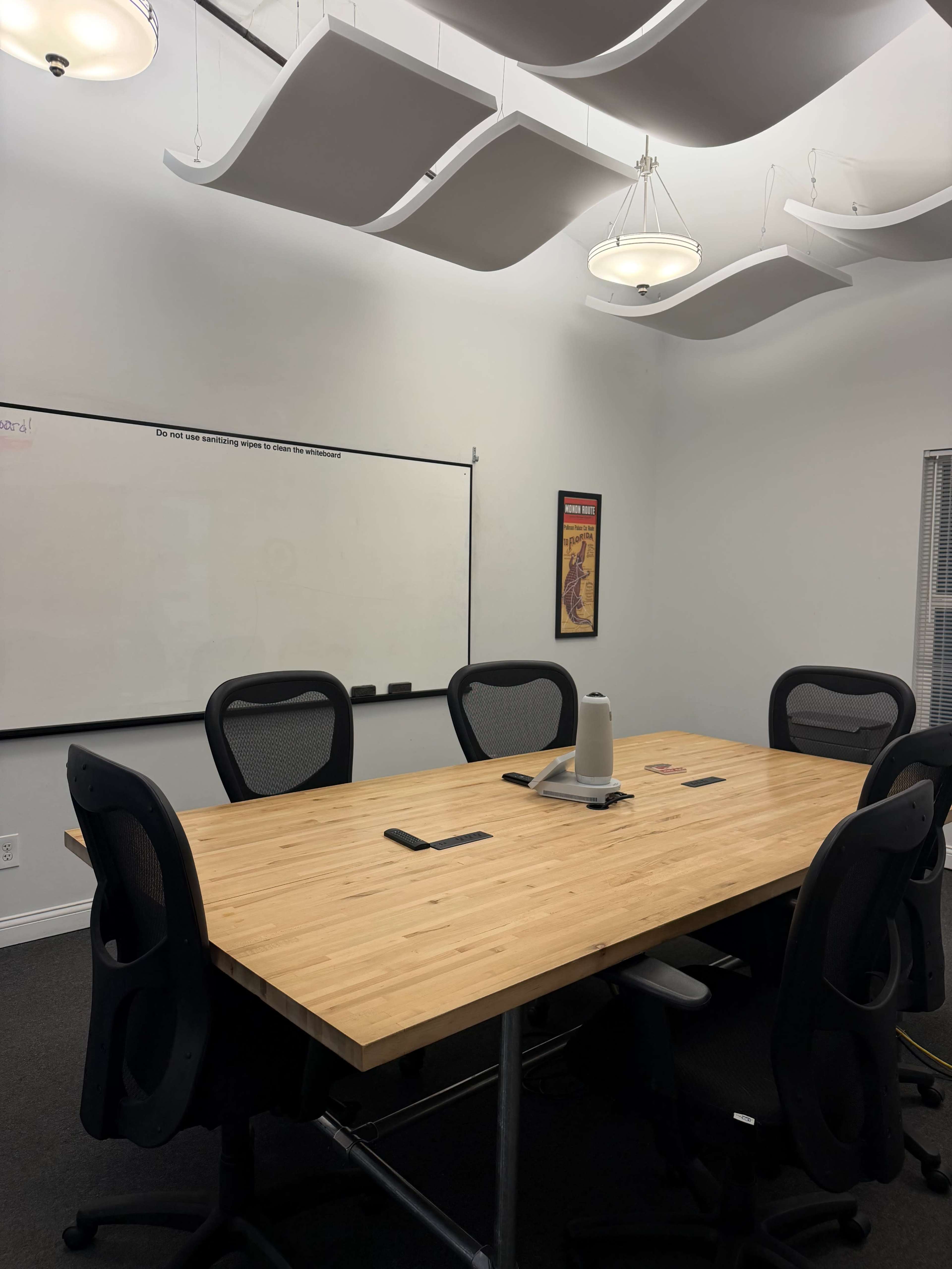 A conference room features a large wooden table surrounded by black chairs, with a whiteboard and decorative wall art in the background.