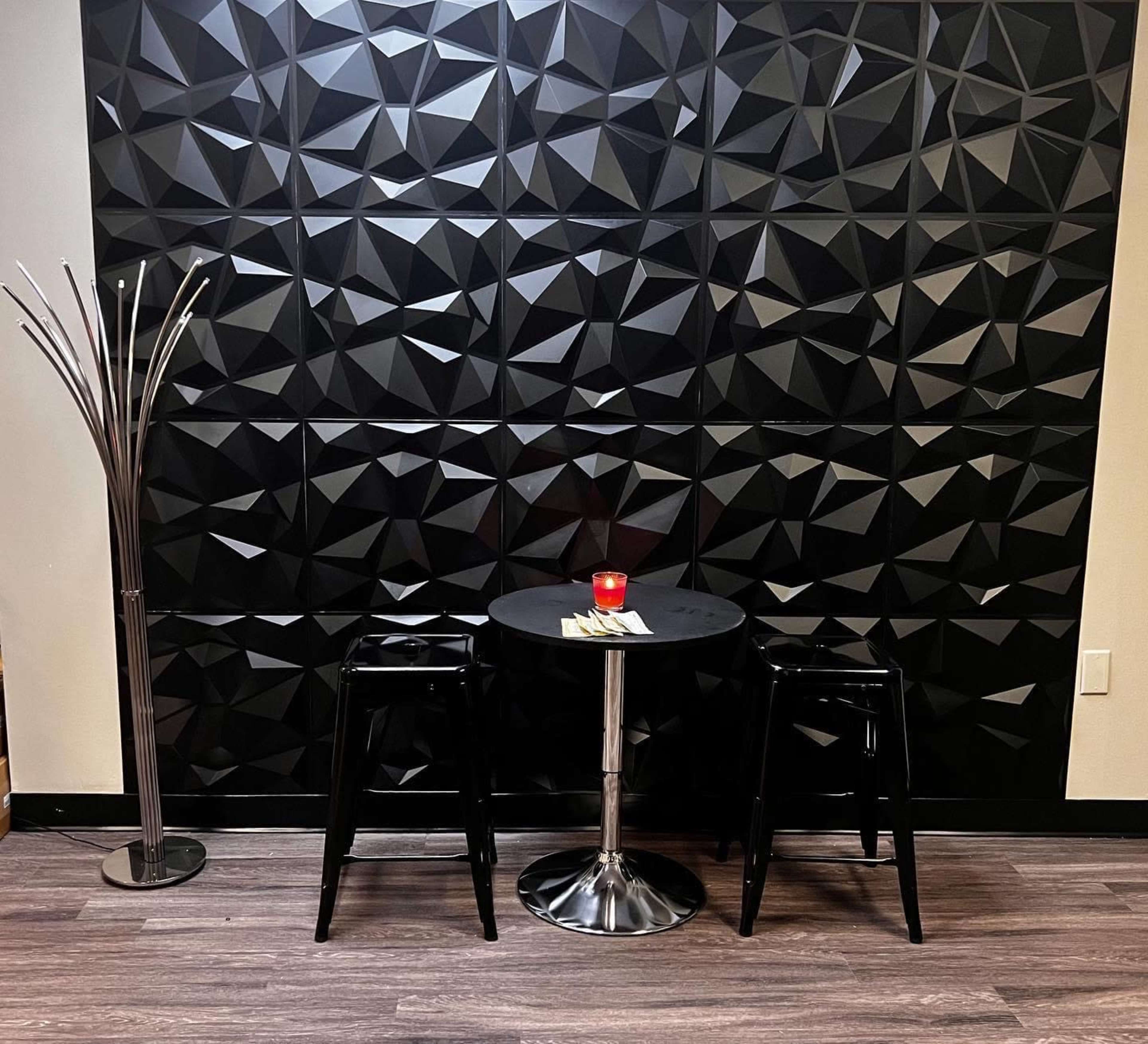 A small round table with two black stools is positioned in front of a textured black wall, beside a modern metallic floor lamp.