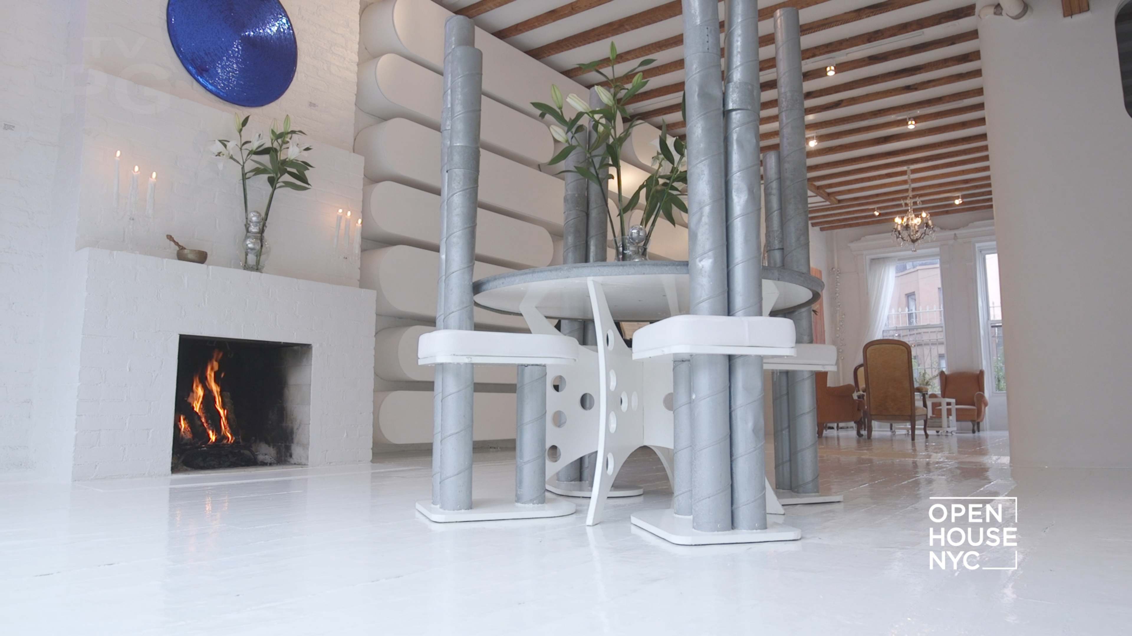 The image shows a modern living space with a unique circular table supported by silver pillars, surrounded by minimalistic decor and a fireplace.