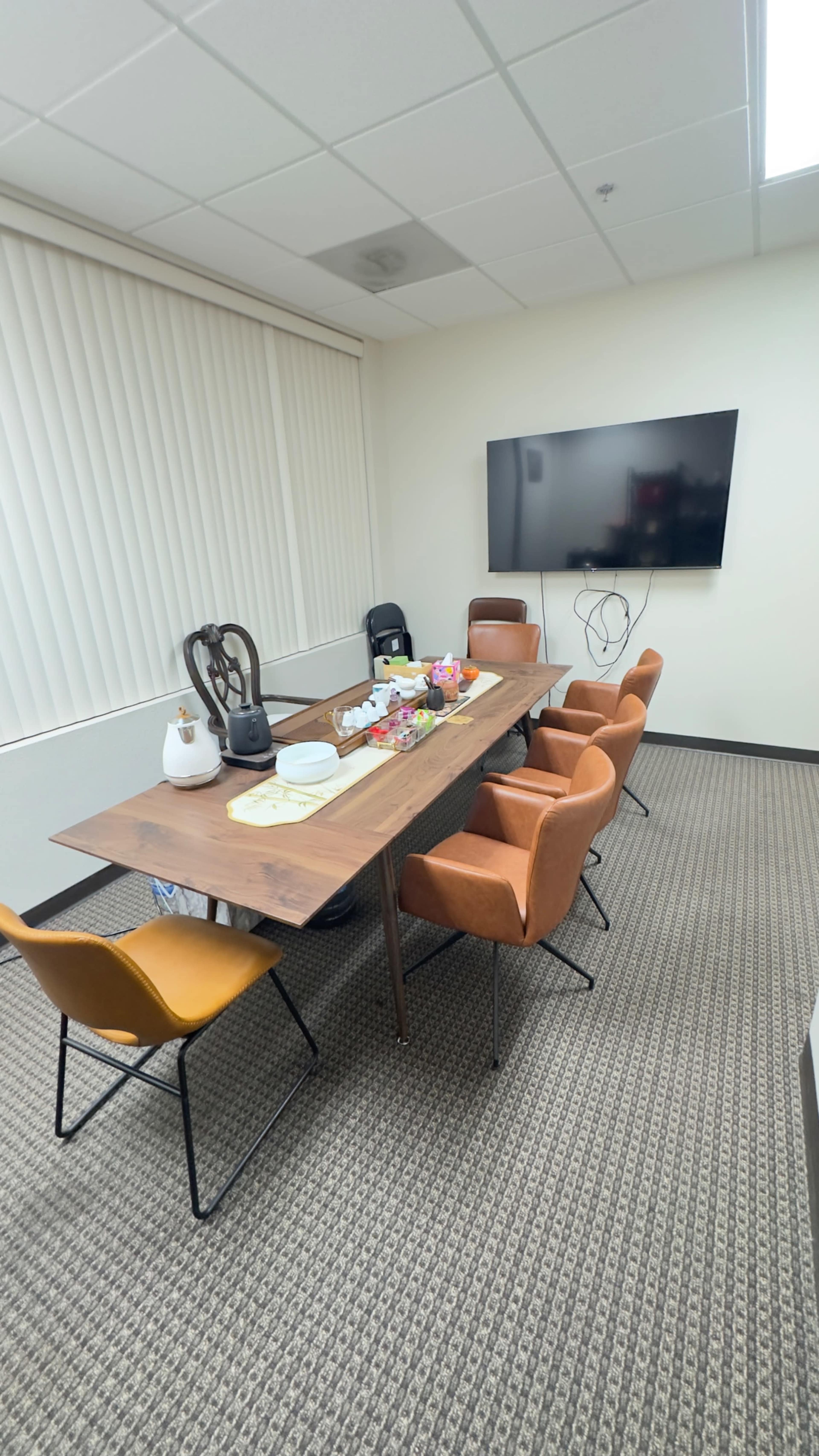 The image shows a conference room with a wooden table surrounded by orange and black chairs, a television mounted on the wall, and snacks arranged on the table.