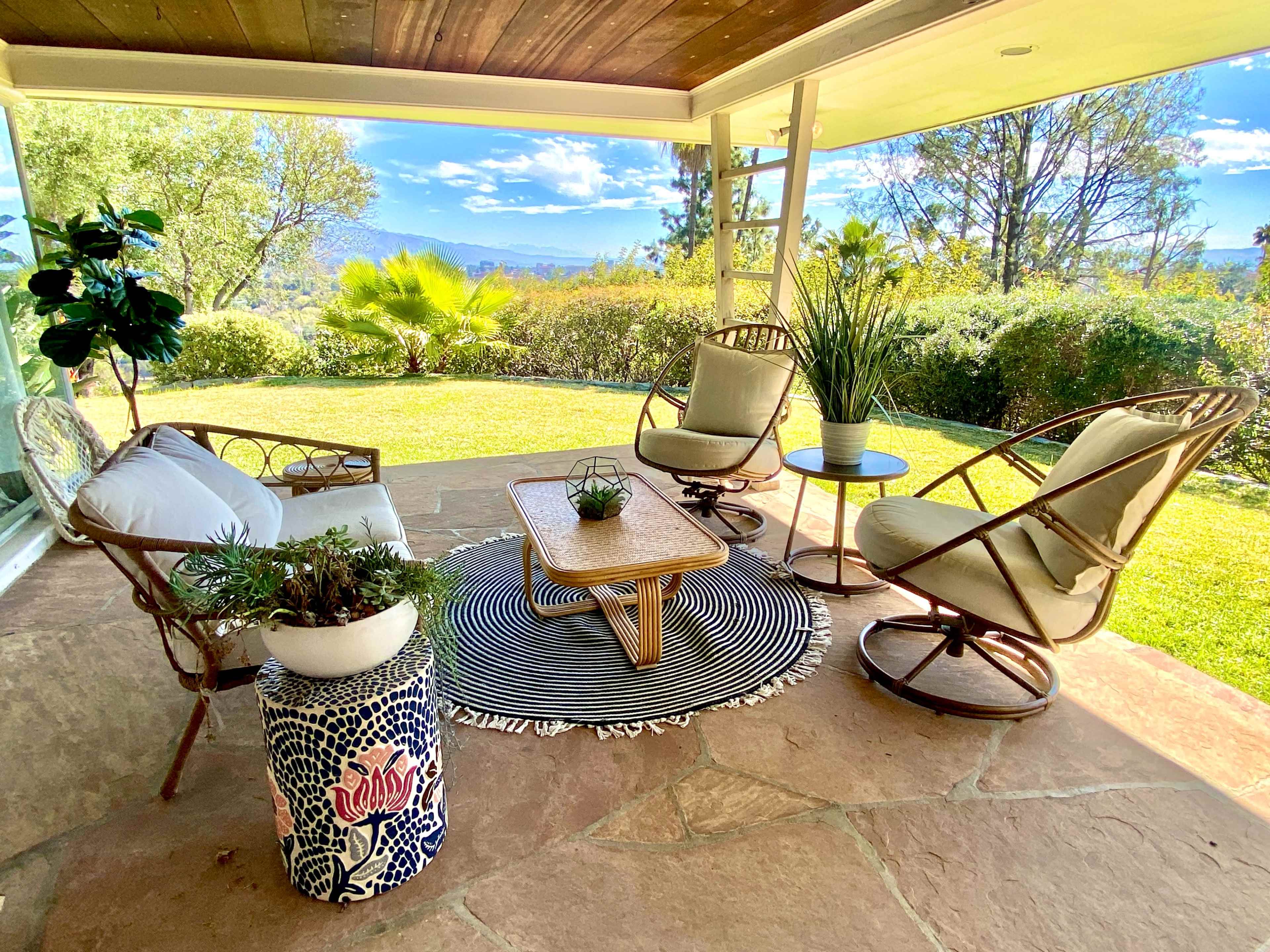 A patio area features two armchairs, a small table, and a striped rug, surrounded by greenery and a scenic view.
