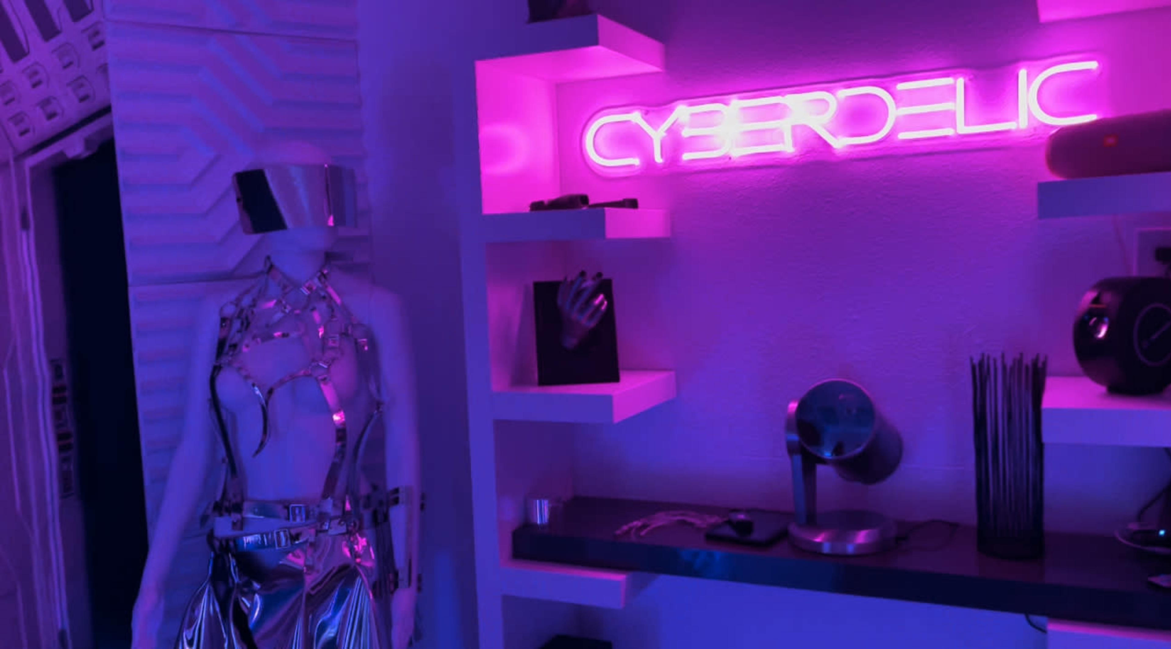 A mannequin dressed in metallic attire stands beside a glowing pink "CYBERDELIC" sign on a shelf with various objects.