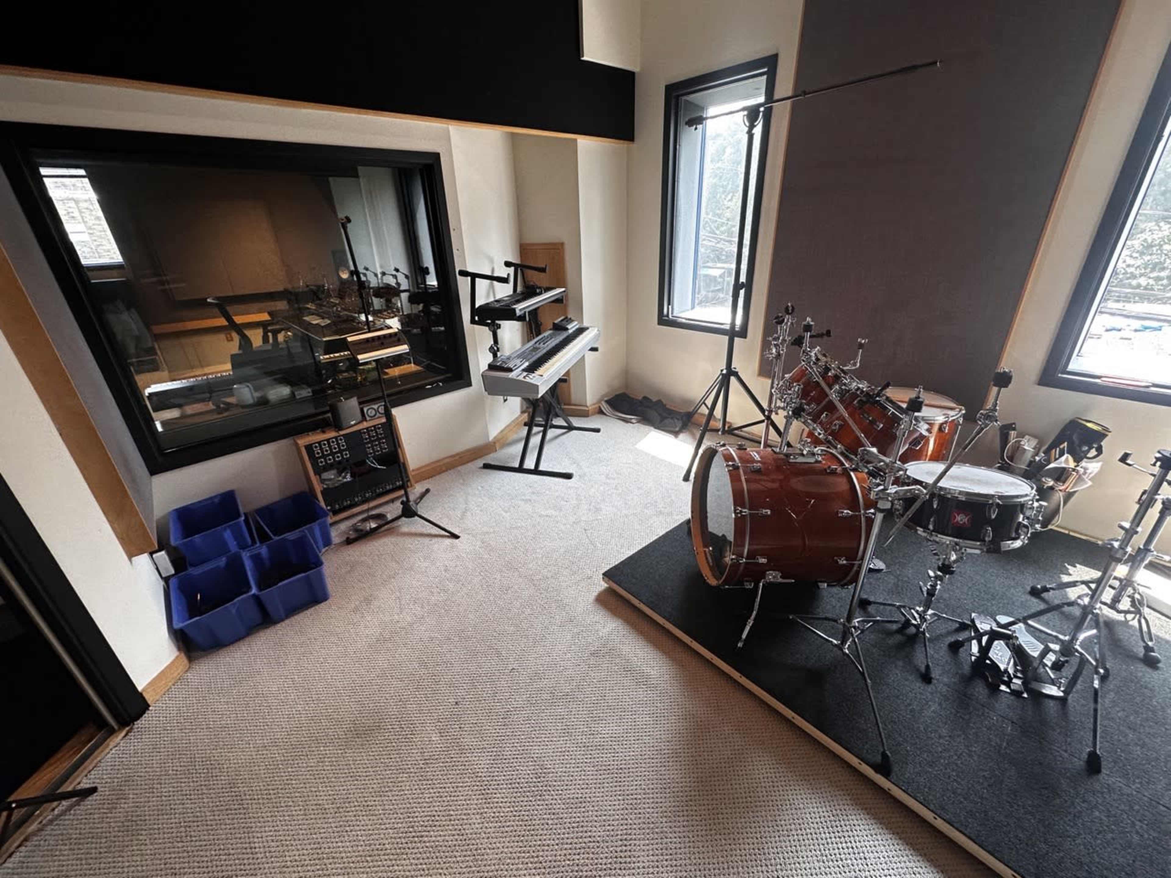 The image shows a music studio with a drum set, keyboard, and an adjoining control room visible through a glass window.
