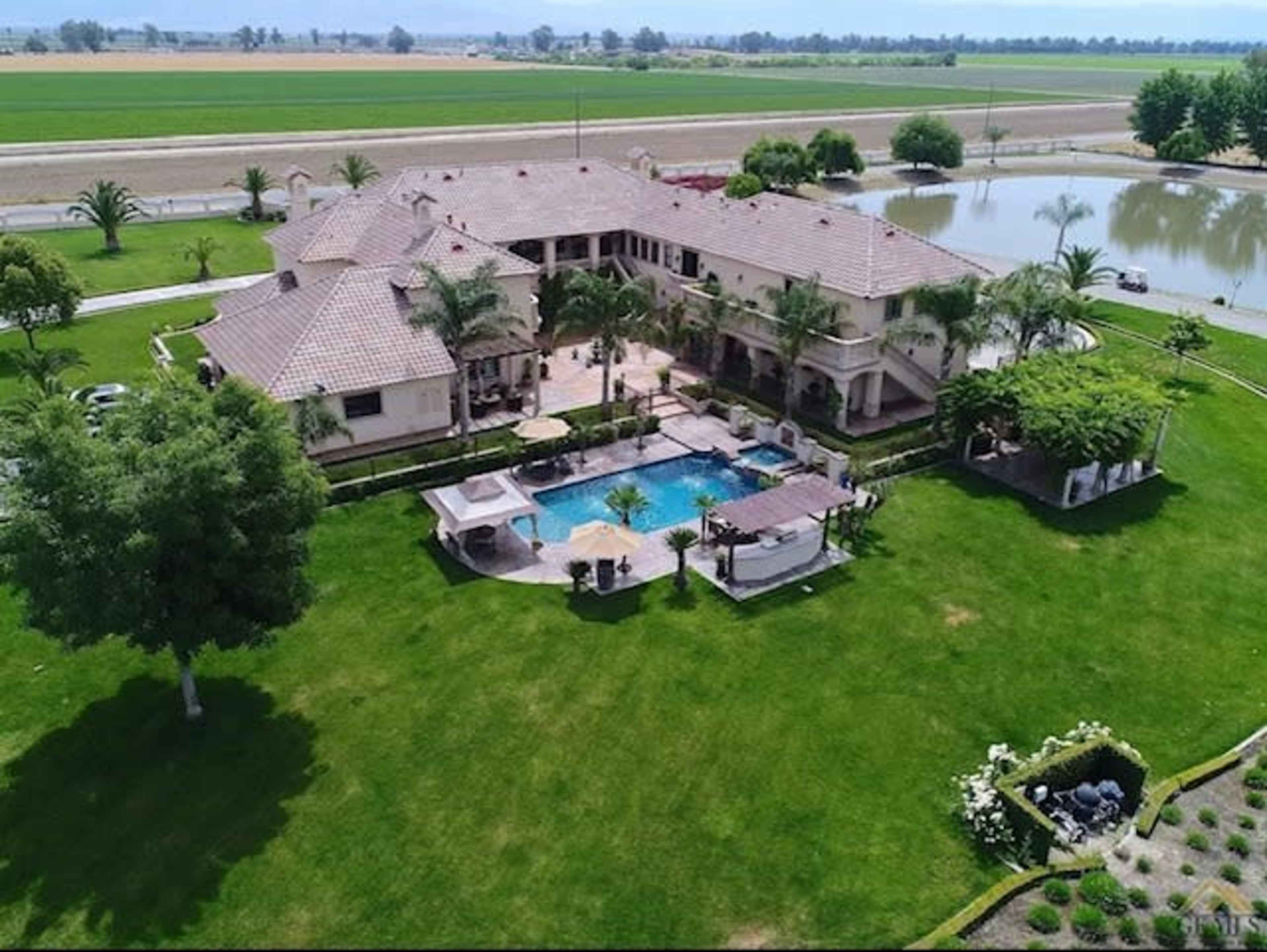 A large, multi-story house surrounded by green lawns, a swimming pool, and a pond, with fields in the background.