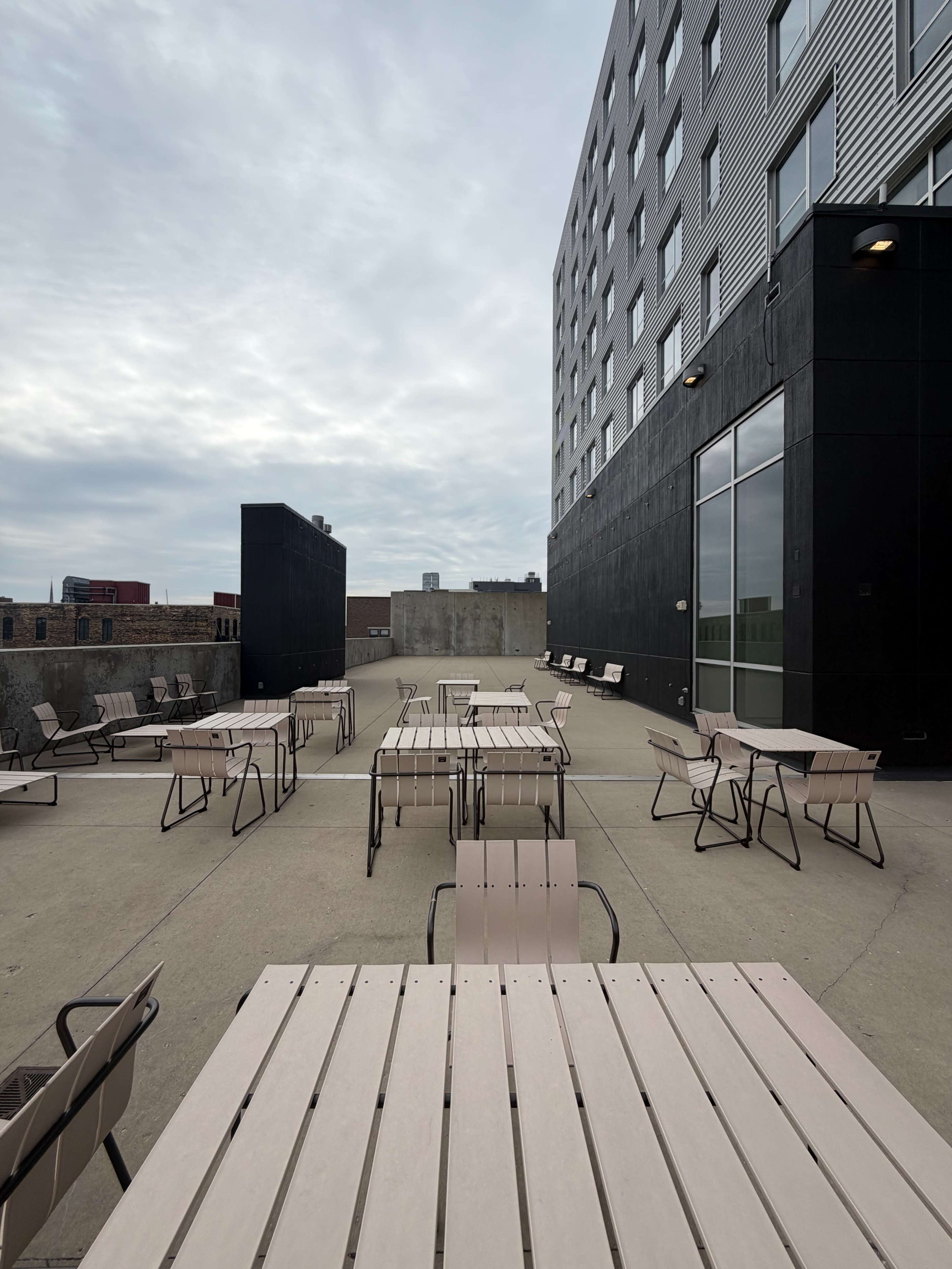 The image shows an outdoor patio area with multiple tables and chairs arranged neatly against a backdrop of a large building.