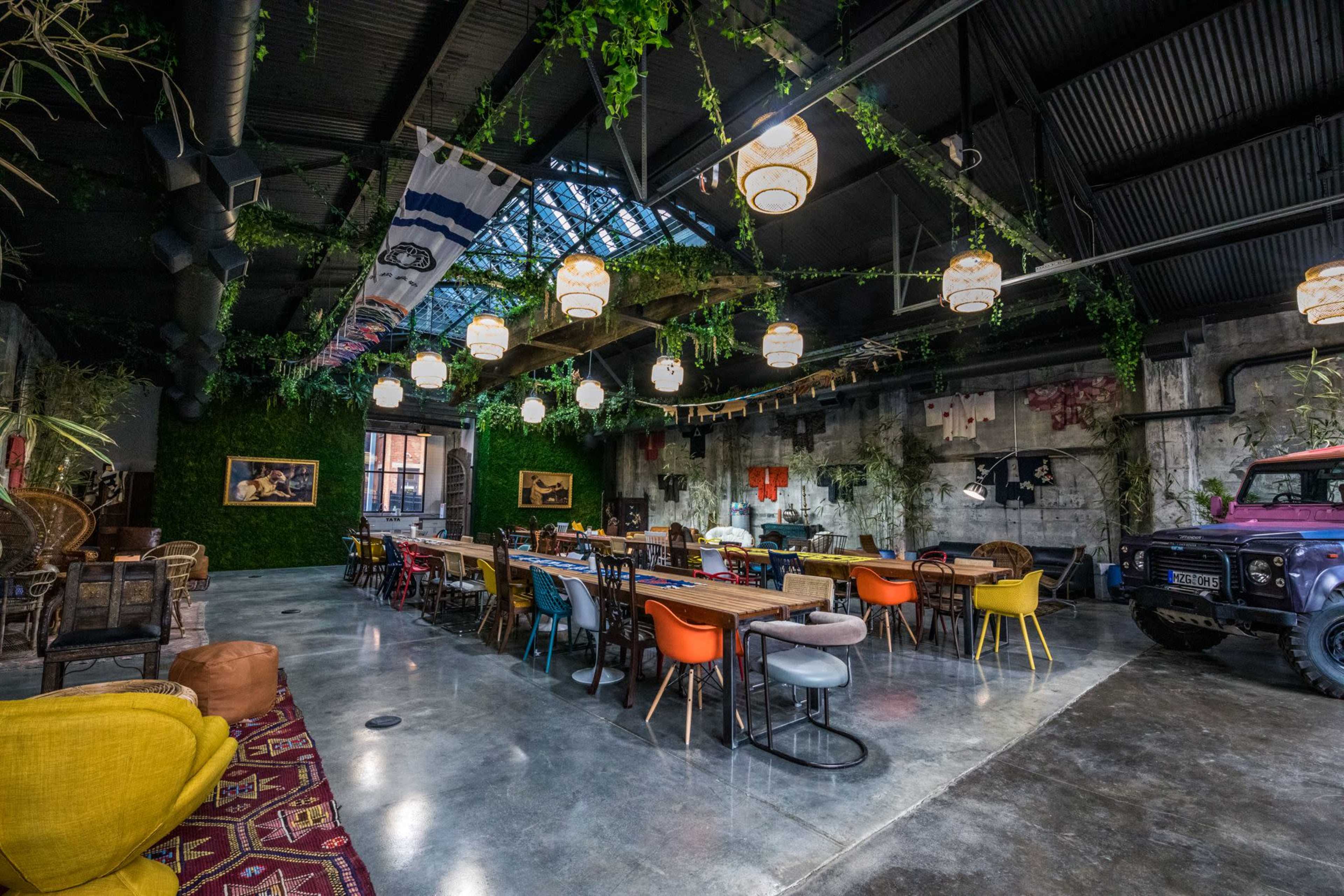 A spacious industrial-style venue with long tables, colorful chairs, and greenery, illuminated by hanging lamps.