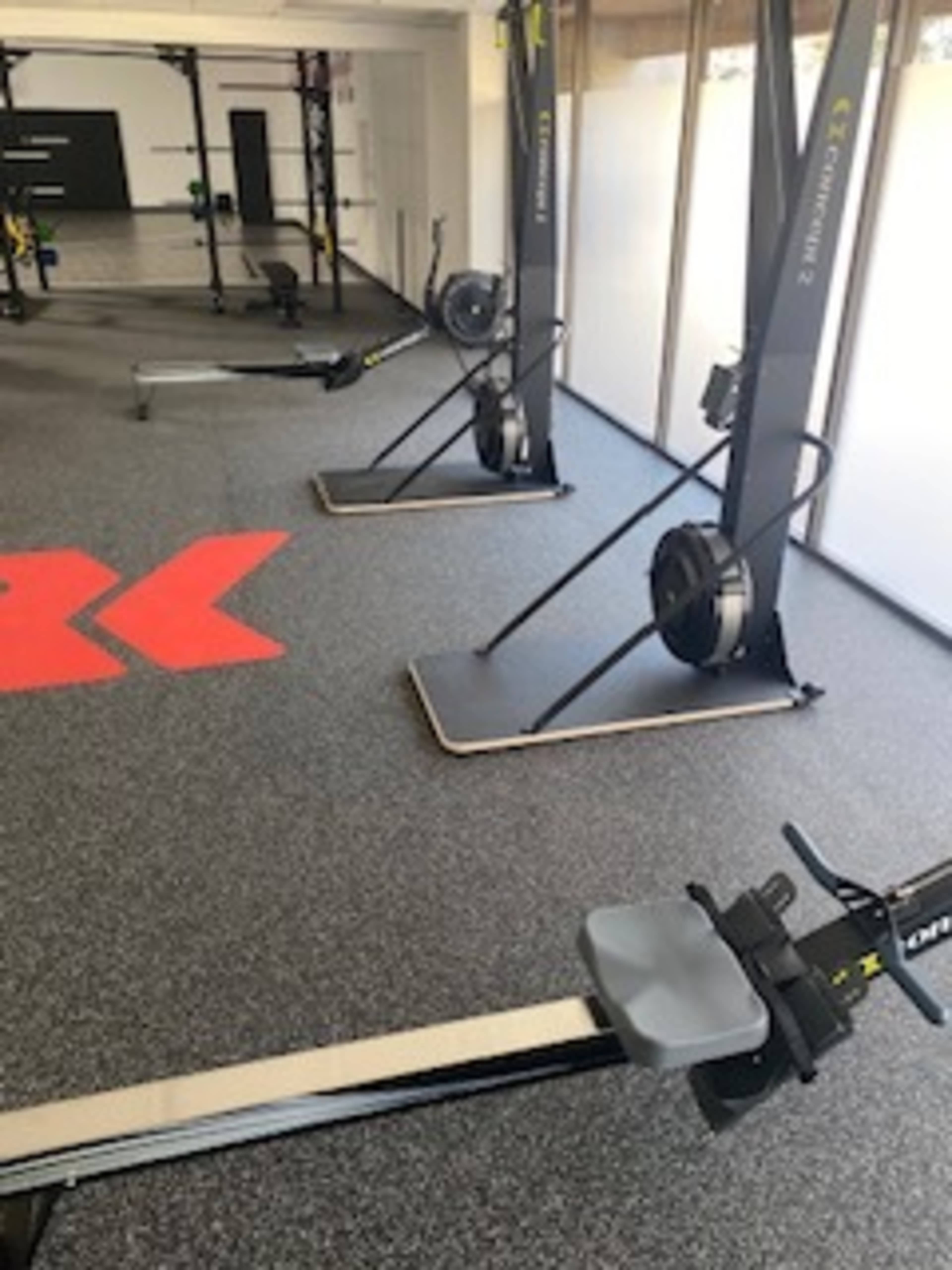 The image shows a fitness studio with rowing machines and exercise equipment arranged on a rubberized floor.