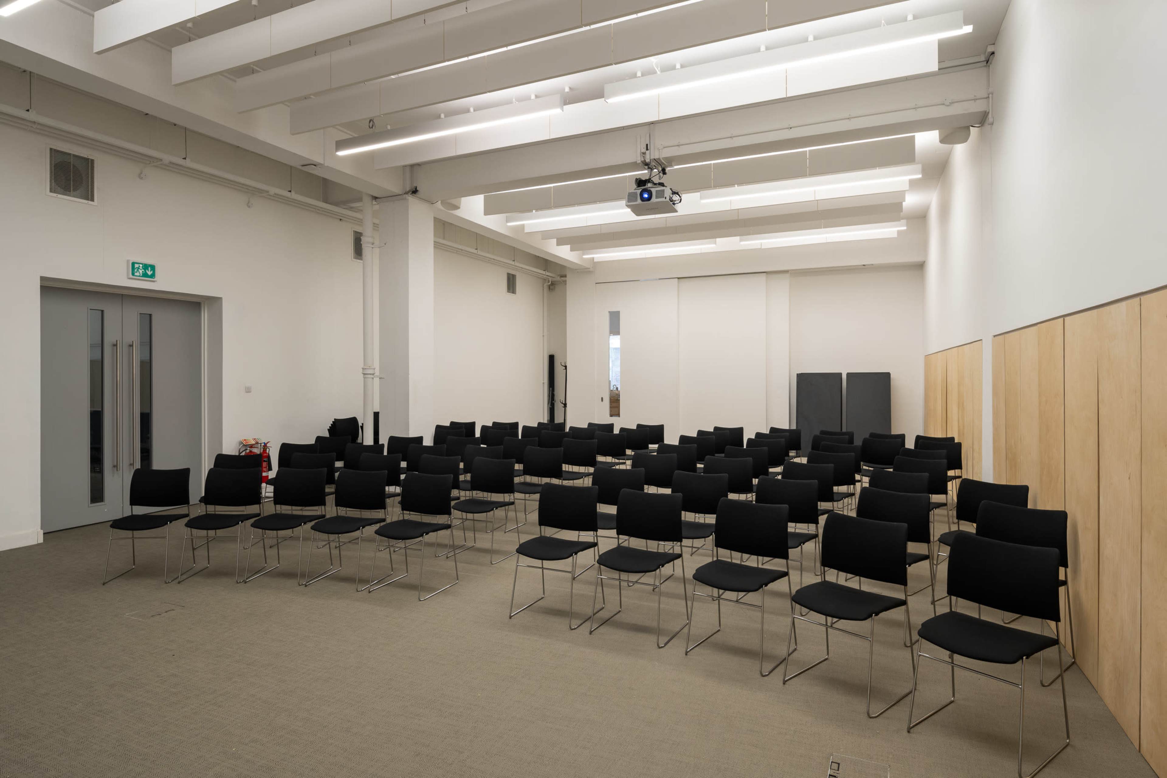 The image shows a sparsely arranged seminar room with rows of black chairs set up facing a projector screen.