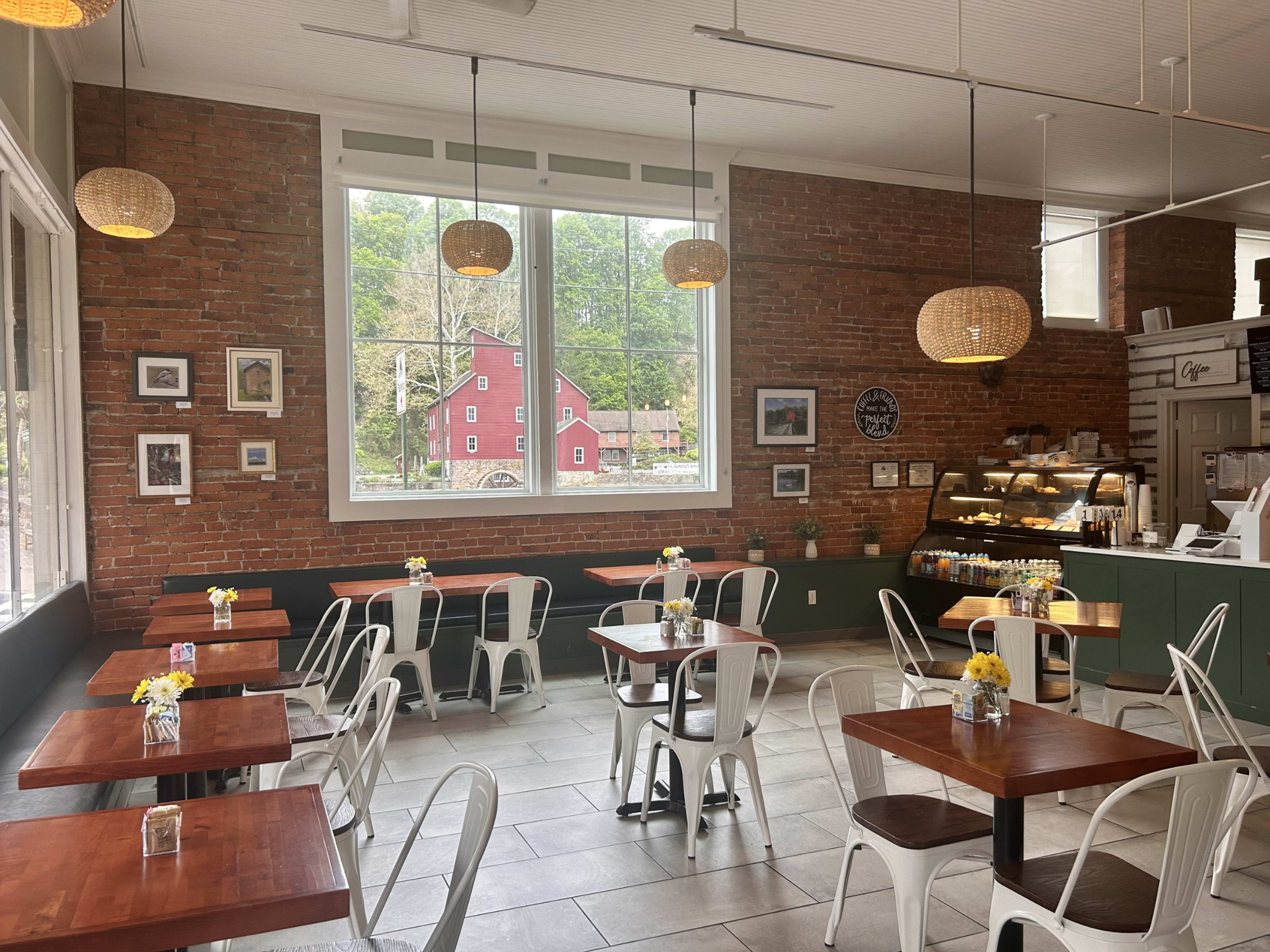 The image shows a cozy café with brick walls, wooden tables, and chairs, featuring large windows that provide natural light and a view of greenery outside.