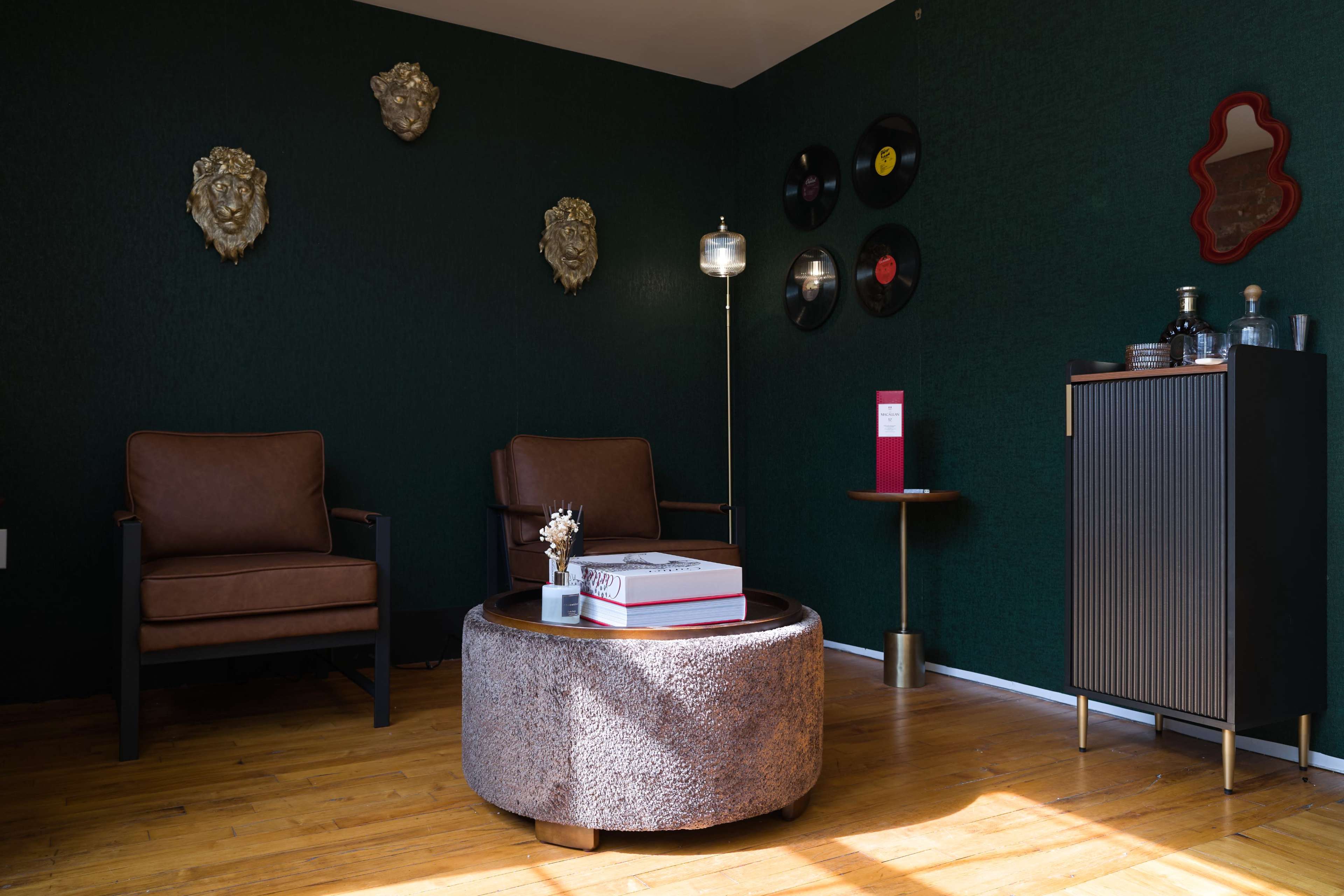 The room features two brown leather chairs, a round ottoman, a standing lamp, and a decorative cabinet against a dark green wall adorned with vintage records and wall art.