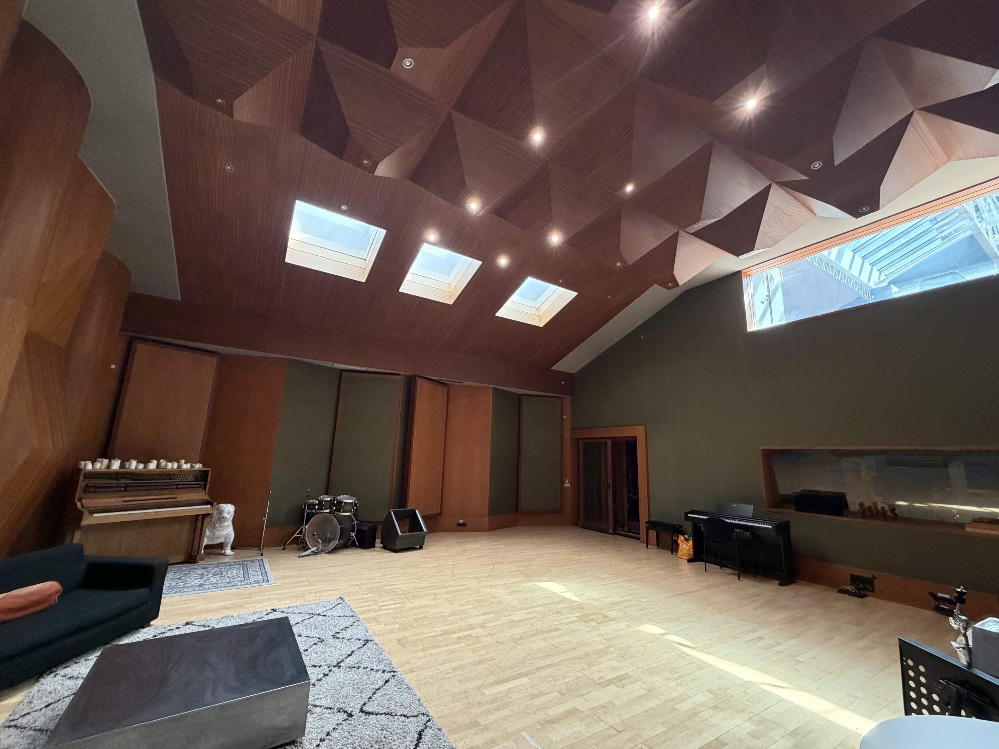 The image shows a spacious music studio with wooden paneling, a high textured ceiling, and skylights, equipped with musical instruments and a sofa.