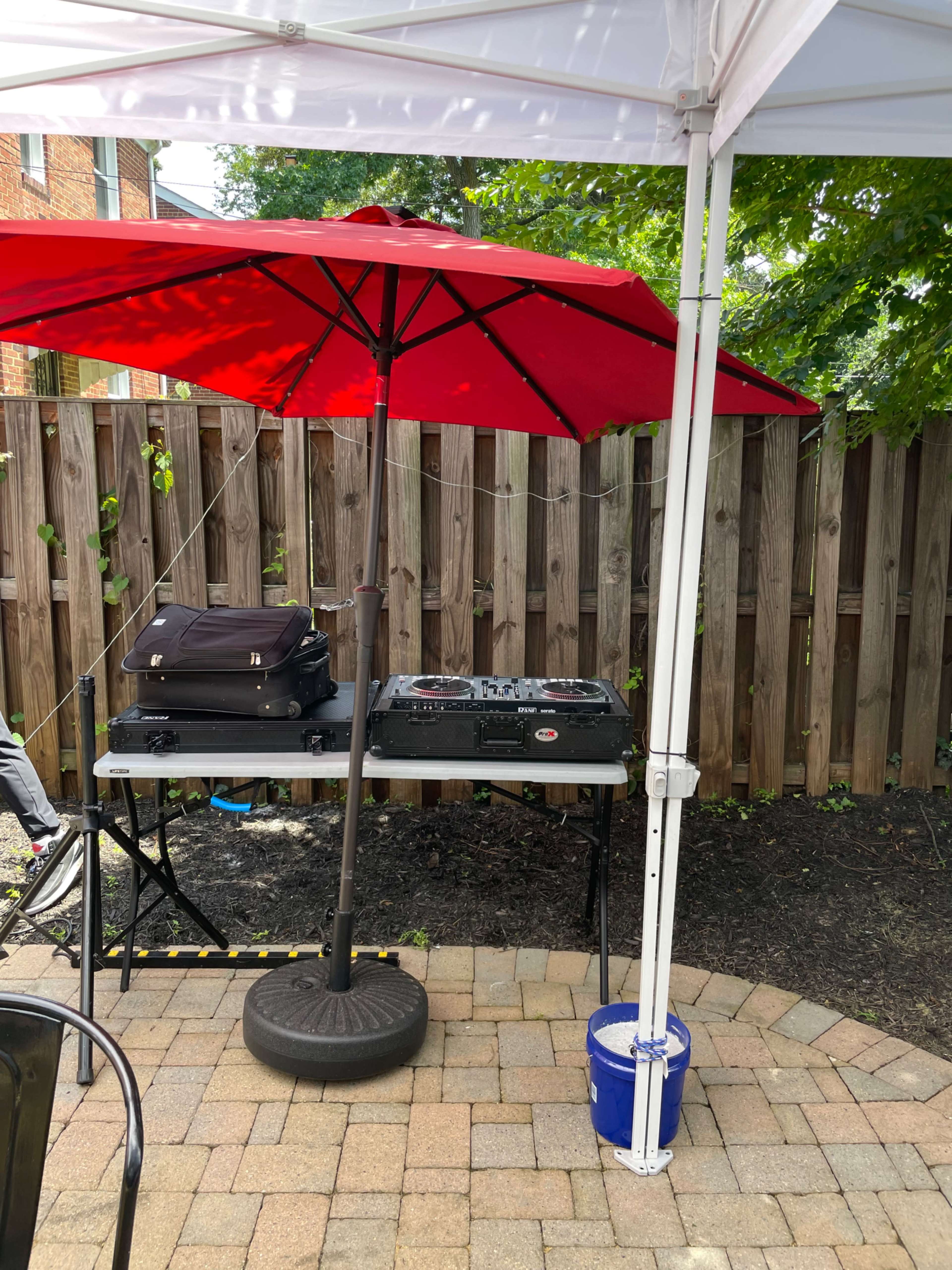 A DJ setup is positioned under a red umbrella on a patio, alongside a black suitcase and a blue bucket.