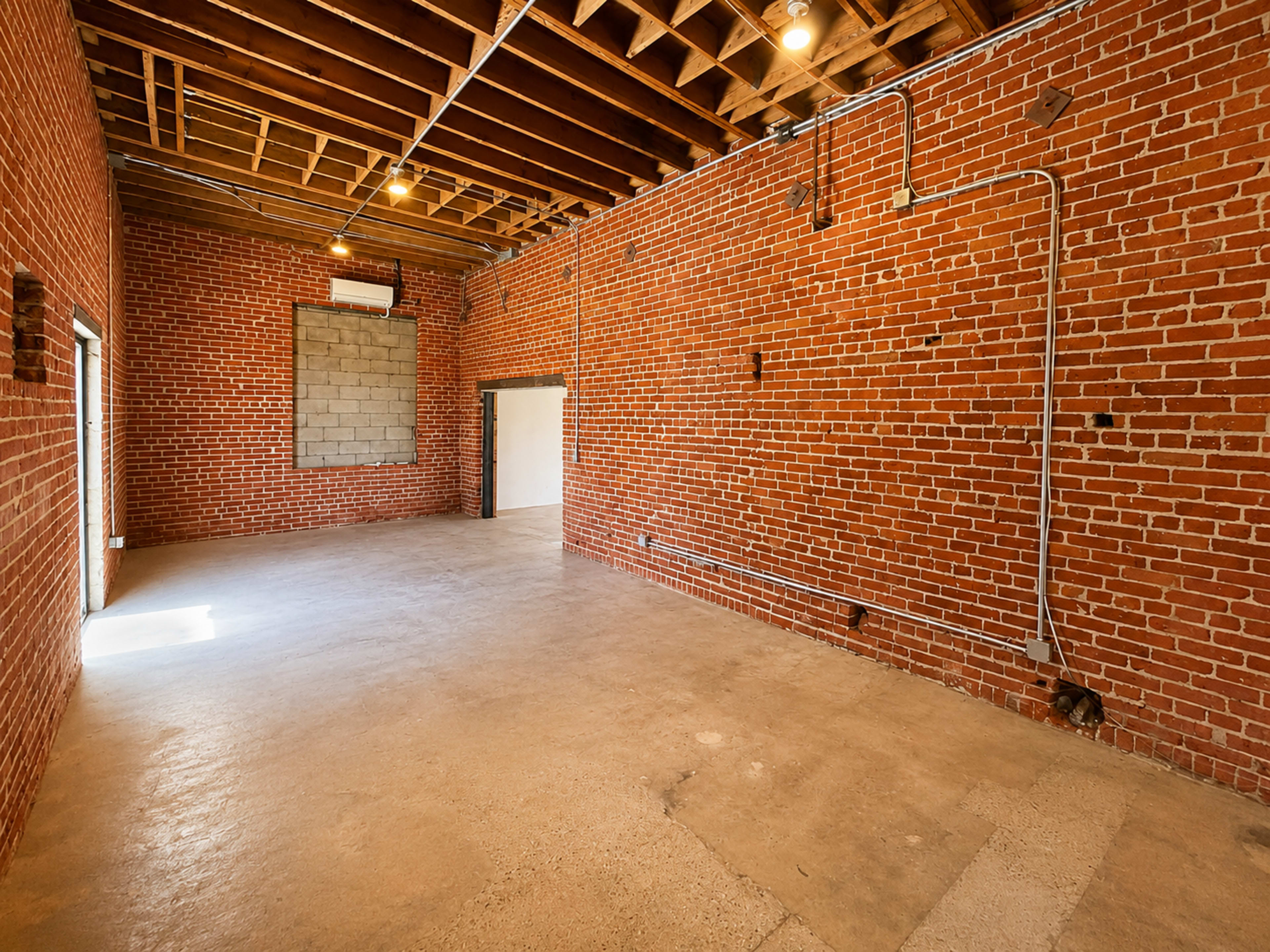 The image shows an empty room with bare brick walls, exposed ceiling beams, and concrete flooring.