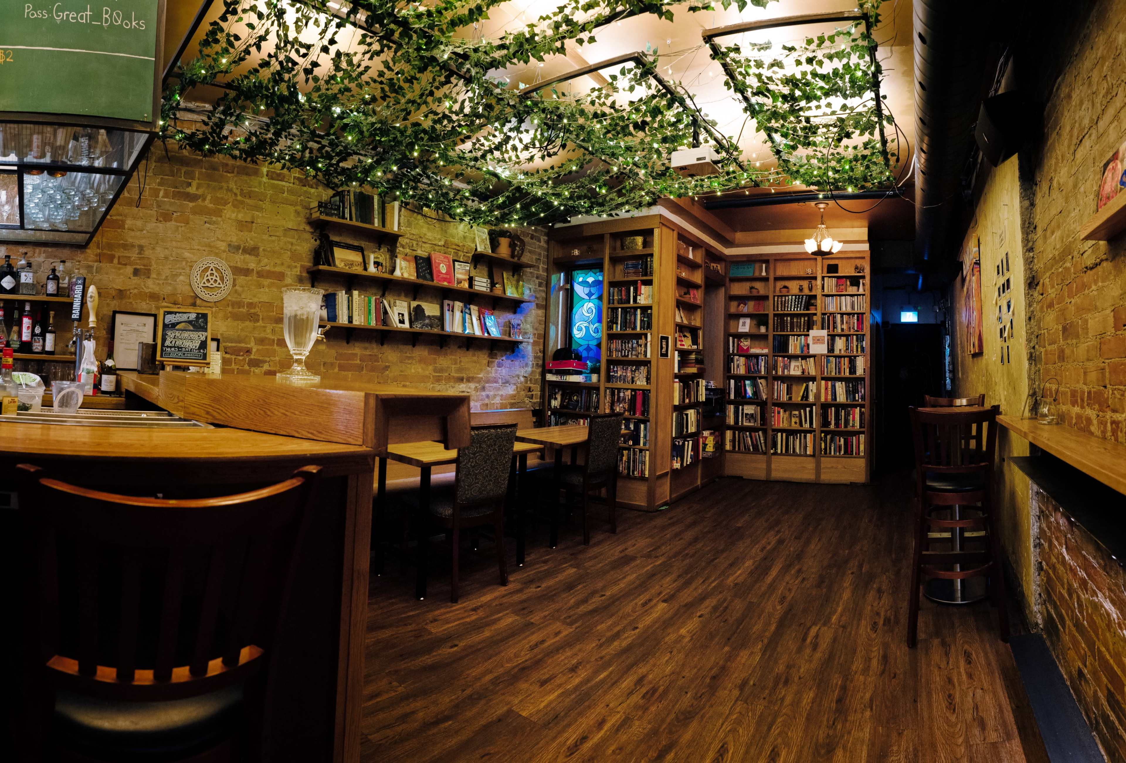 A cozy café features wooden tables, a bookshelf lined with books, and greenery hanging from the ceiling.