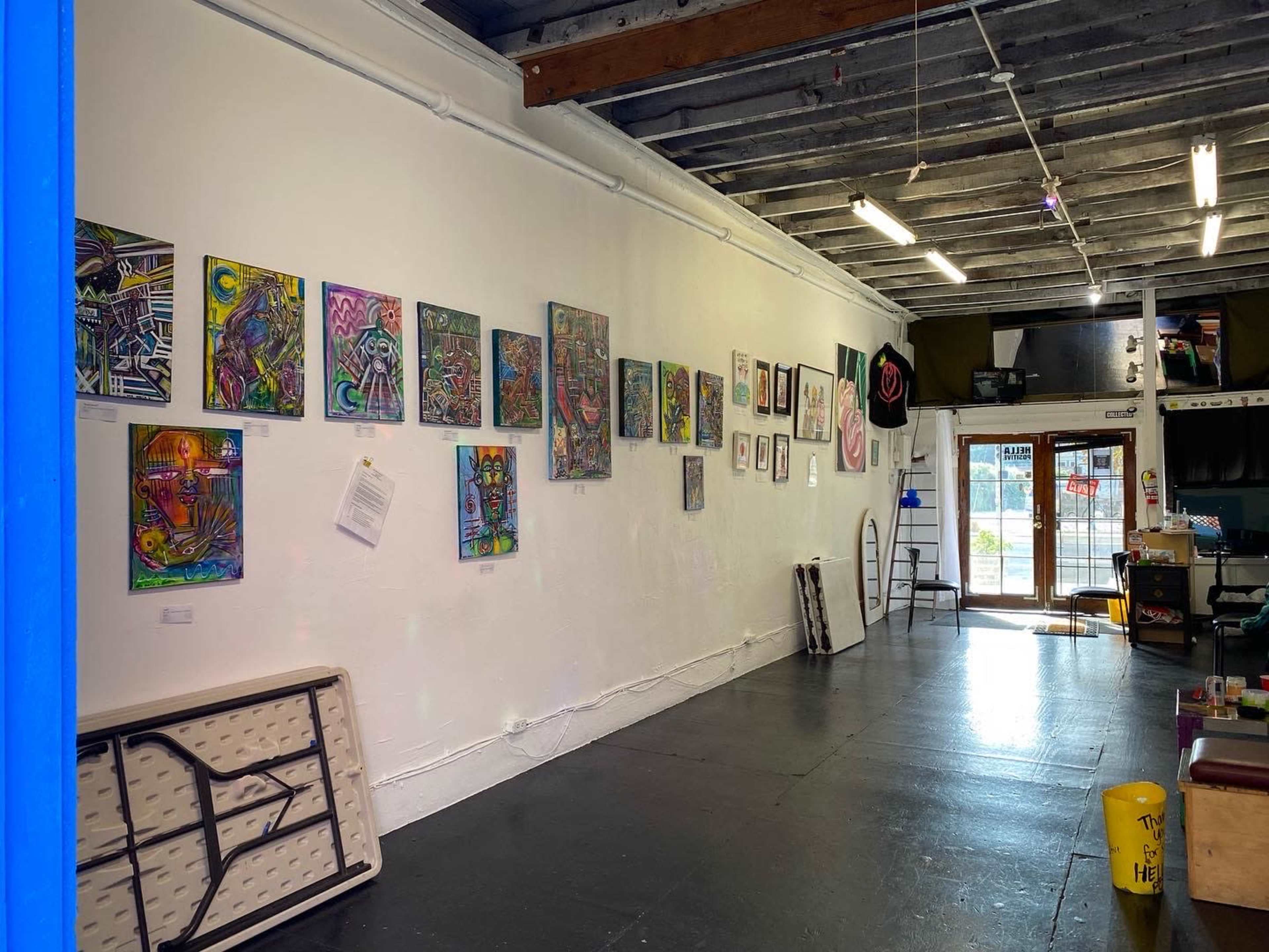 The image shows an art gallery featuring colorful paintings displayed on a white wall, with a folding table and a ladder visible in the interior space.
