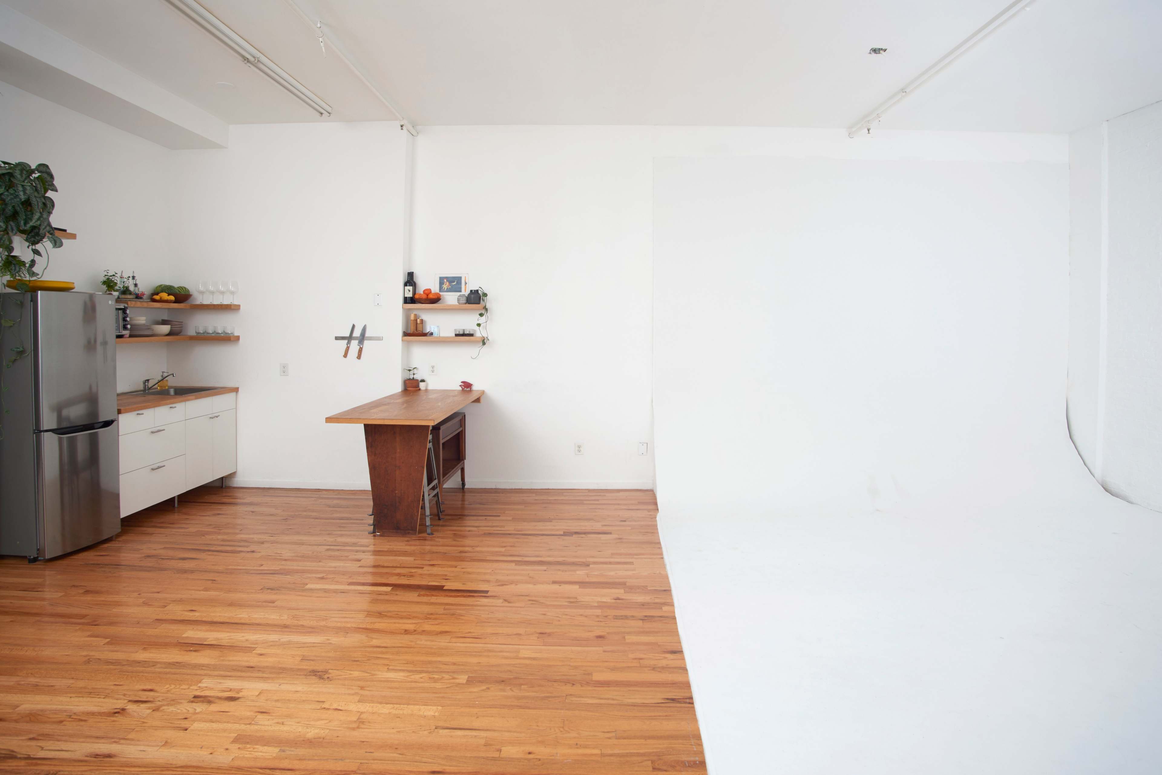 A bright, spacious studio features wooden flooring, a simple kitchen setup with a refrigerator, and a large, empty white backdrop.