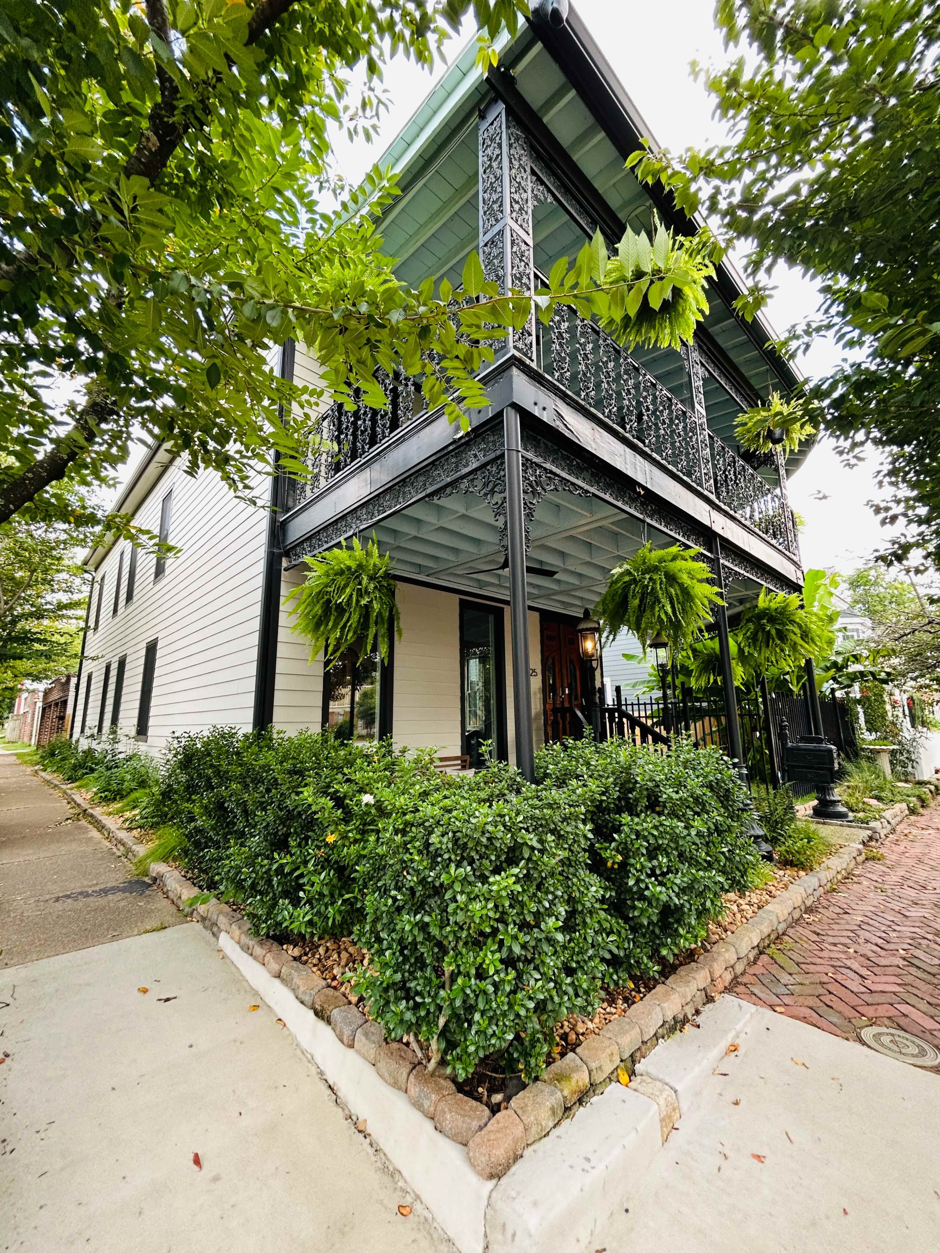 A two-story house with a balcony and decorative railings is surrounded by lush greenery and a brick pathway.