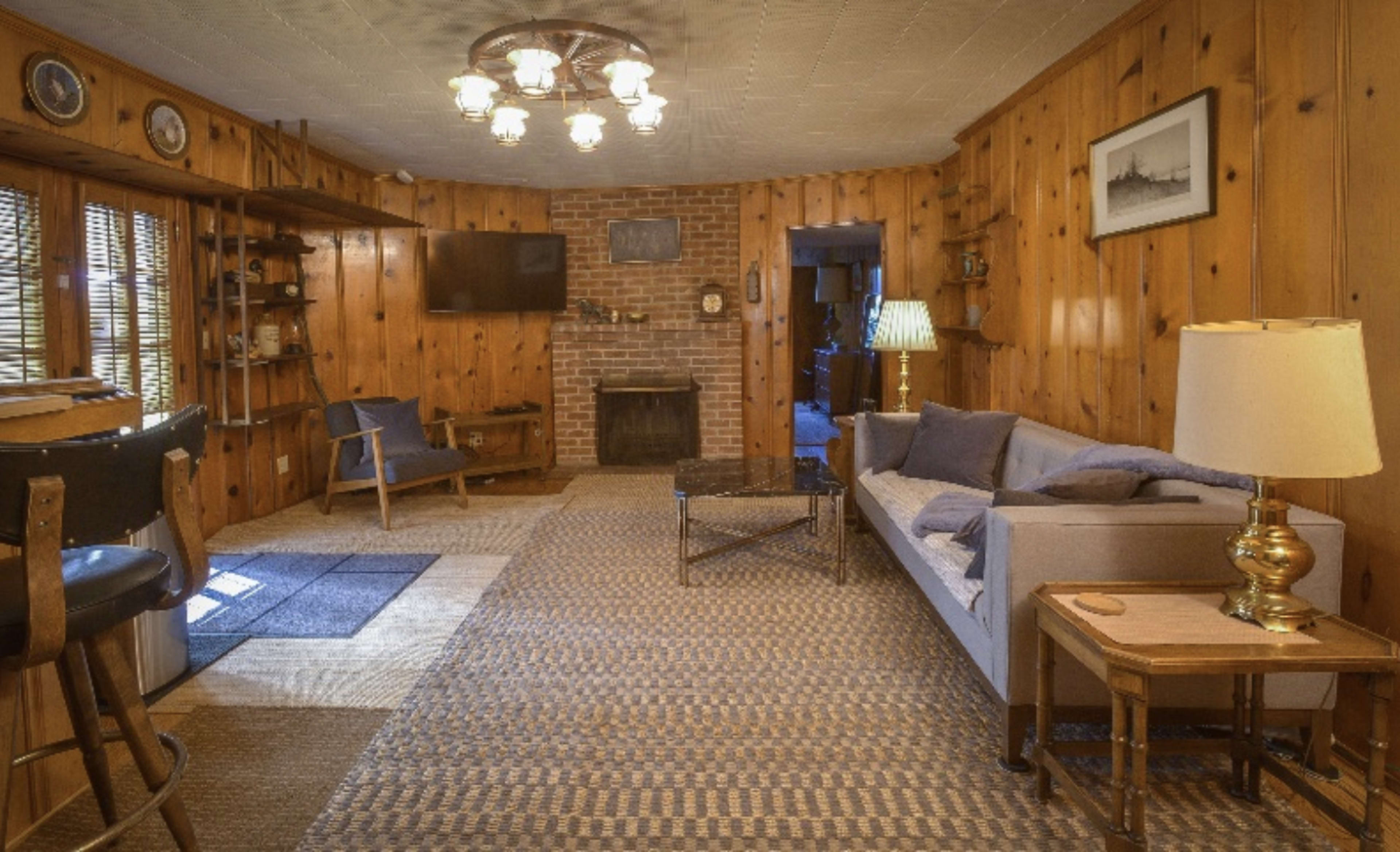 A cozy living room with wooden walls, a brick fireplace, and a mix of furniture including a couch, chairs, and a table.