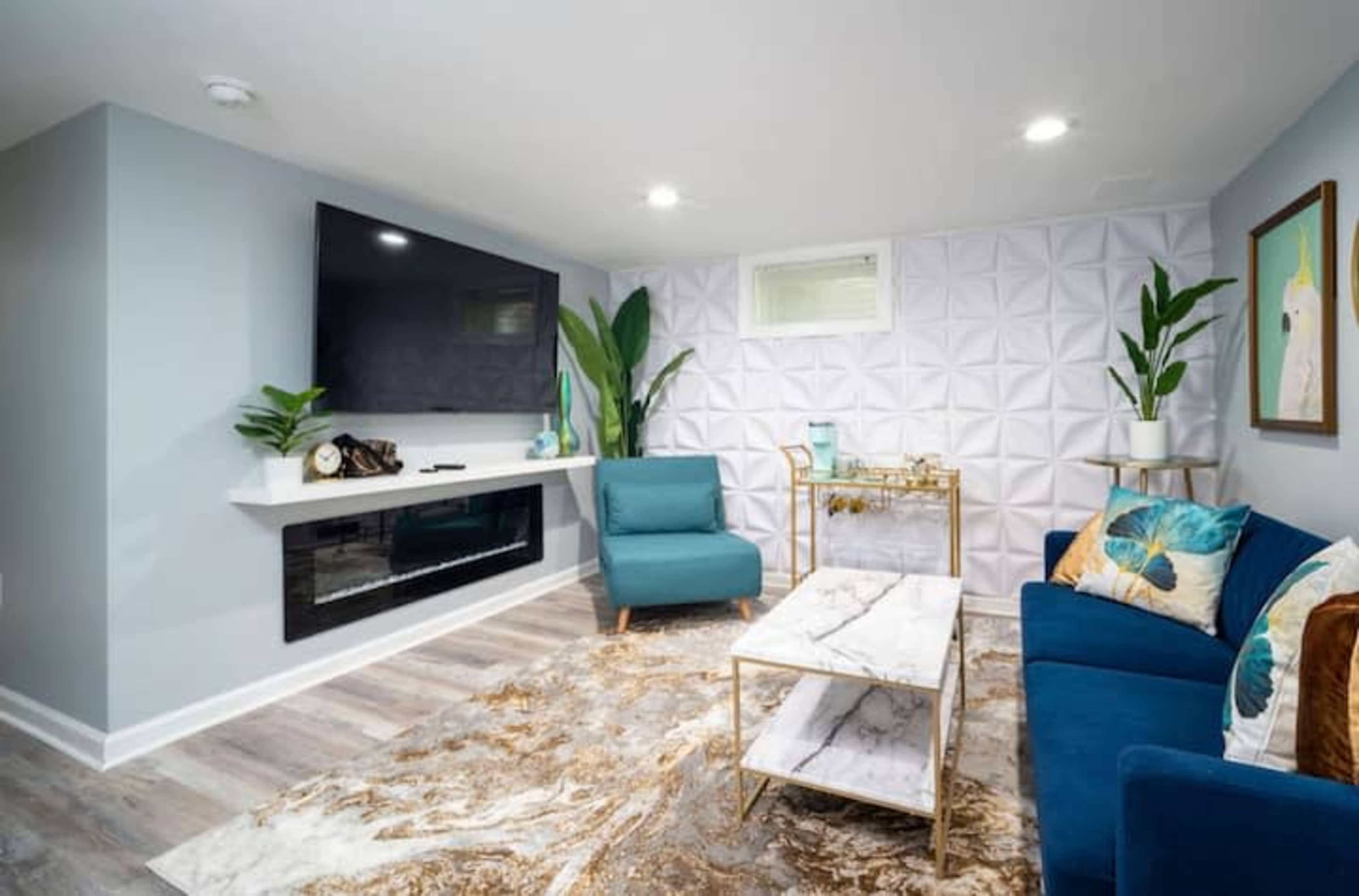 A modern living room with a large flat-screen TV mounted on the wall, a white textured backdrop, and blue seating arranged around a marble-topped coffee table.
