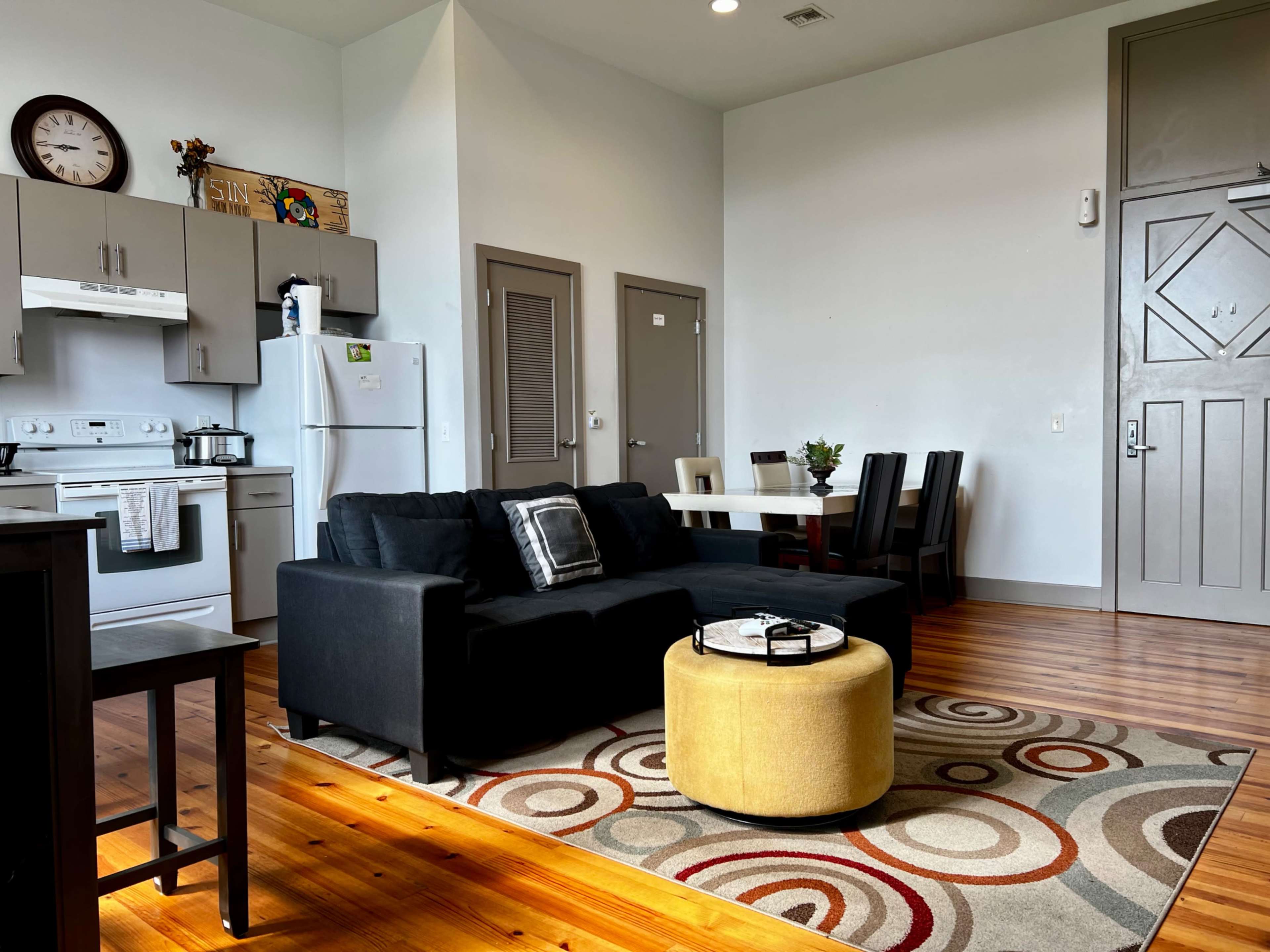 The image shows a modern living space with a black sofa, a round yellow ottoman, a dining table with chairs, and a kitchen area in the background.