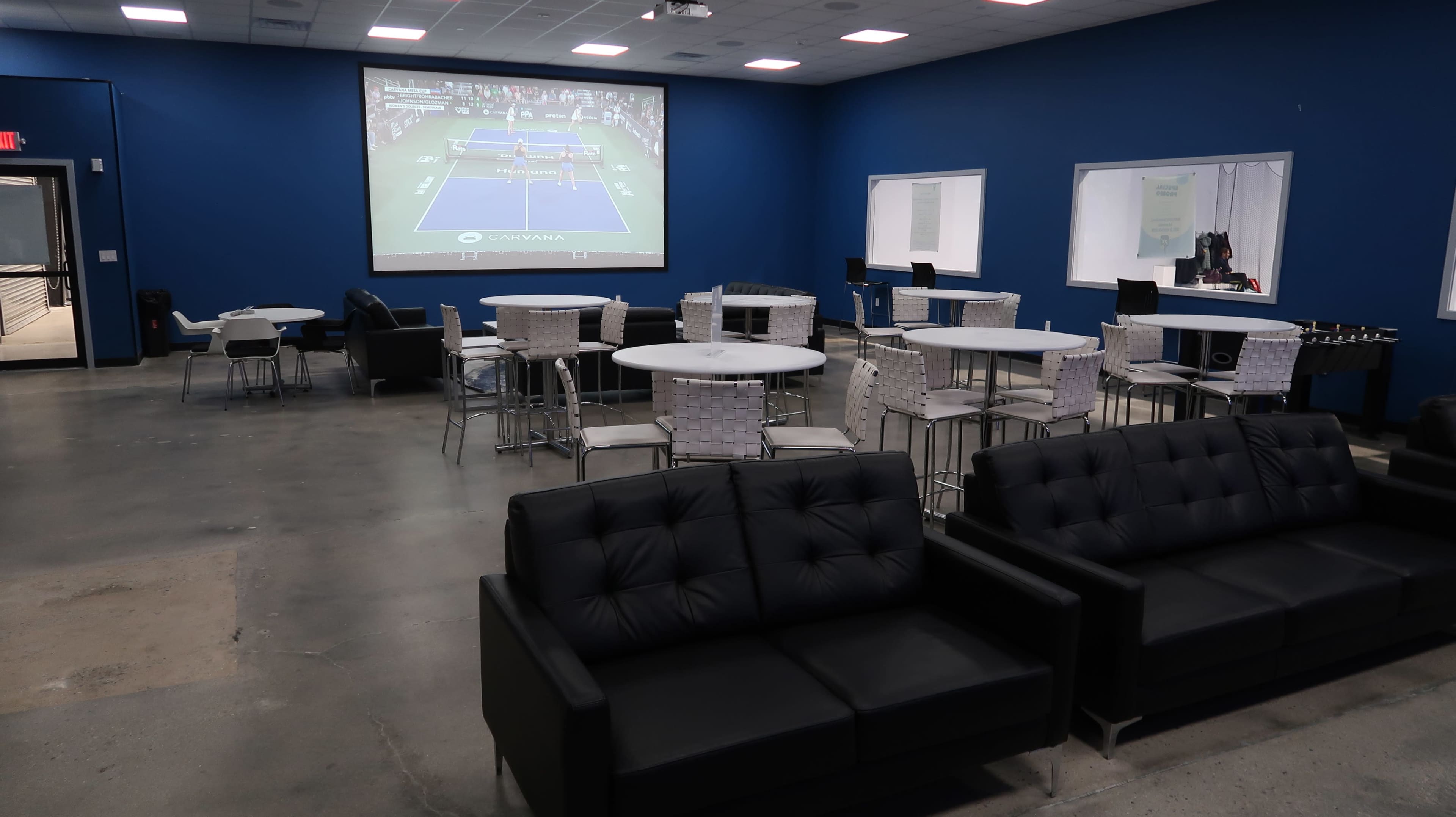 The image shows a recreational room with black sofas, tables, and chairs, and a large screen displaying a tennis match on a blue wall.