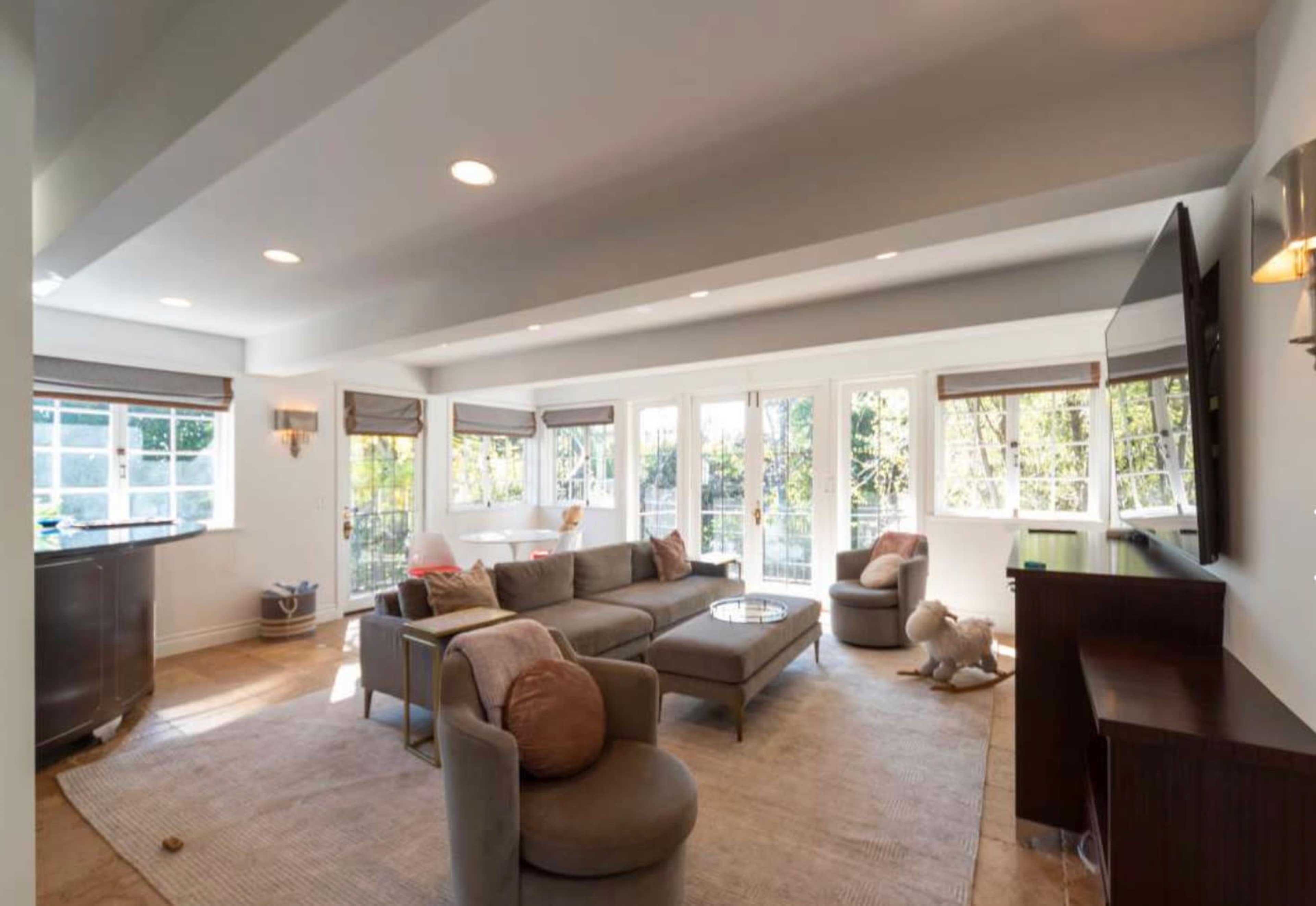 The image shows a modern living room with large windows, a plush sectional sofa, and a television on a wooden stand.