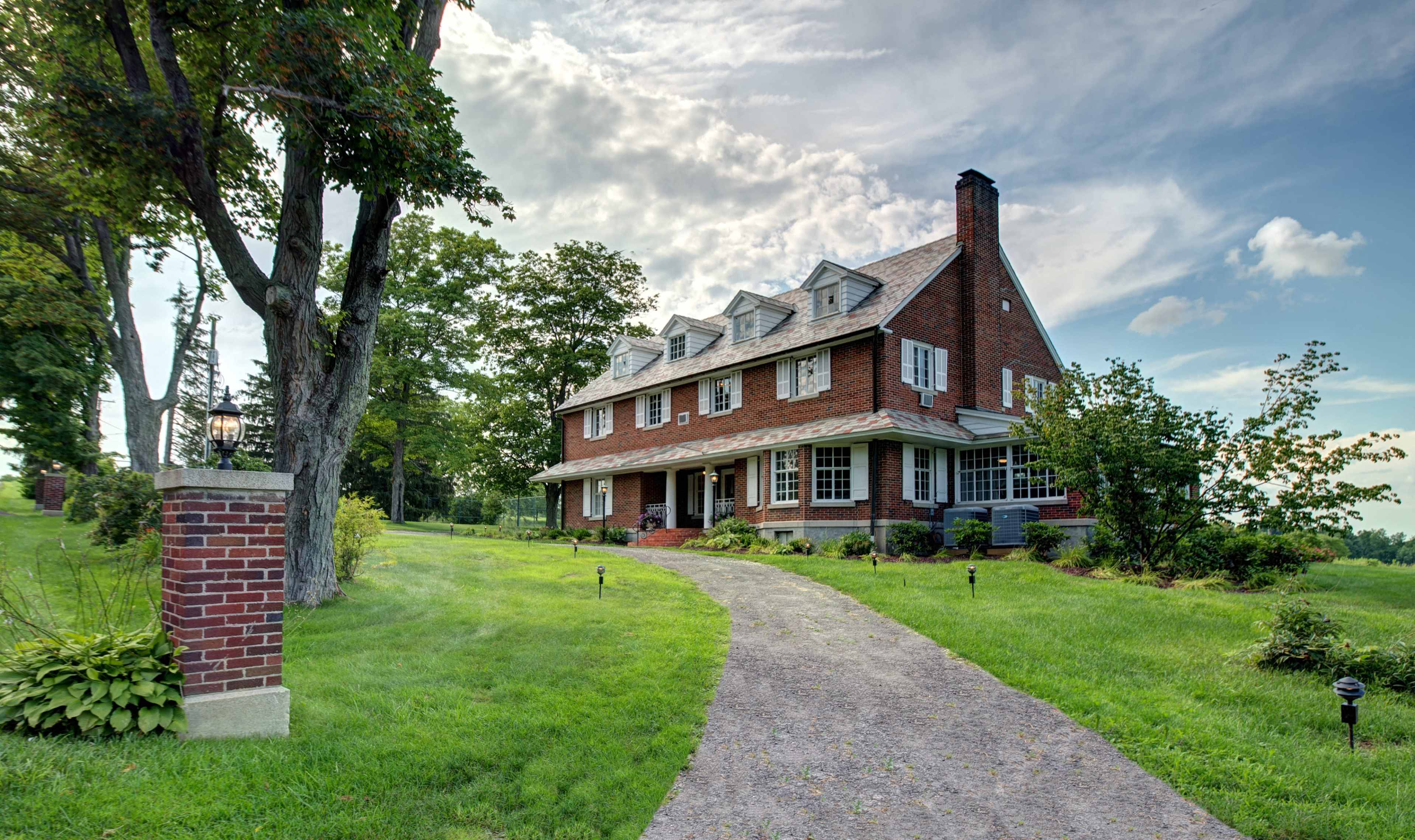 A large brick house with multiple gables sits on a grassy lot, framed by trees and a gravel driveway.