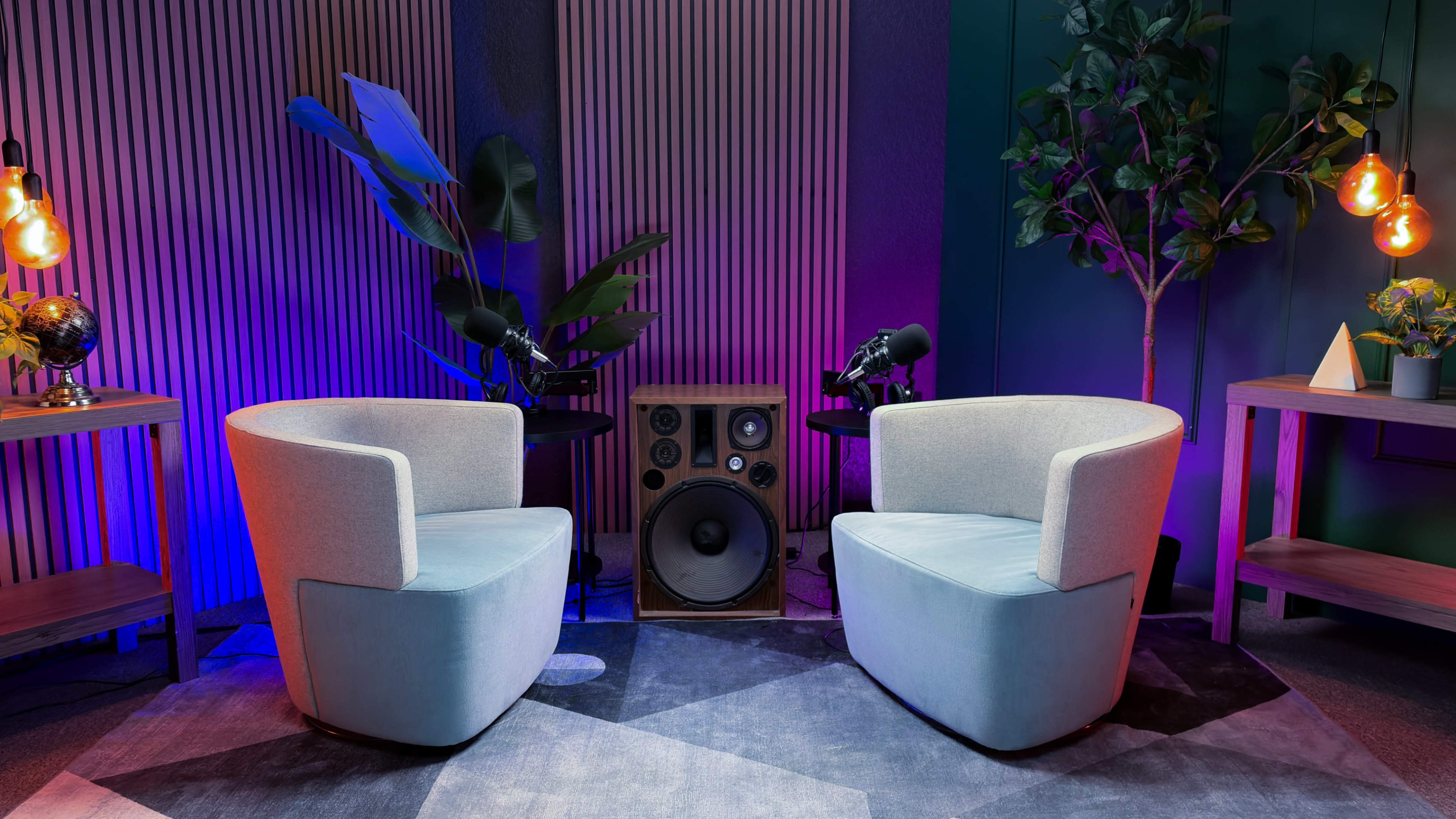 The image shows two modern, light-colored chairs facing each other in a room with purple and blue lighting, alongside a speaker and two small tables with microphones.