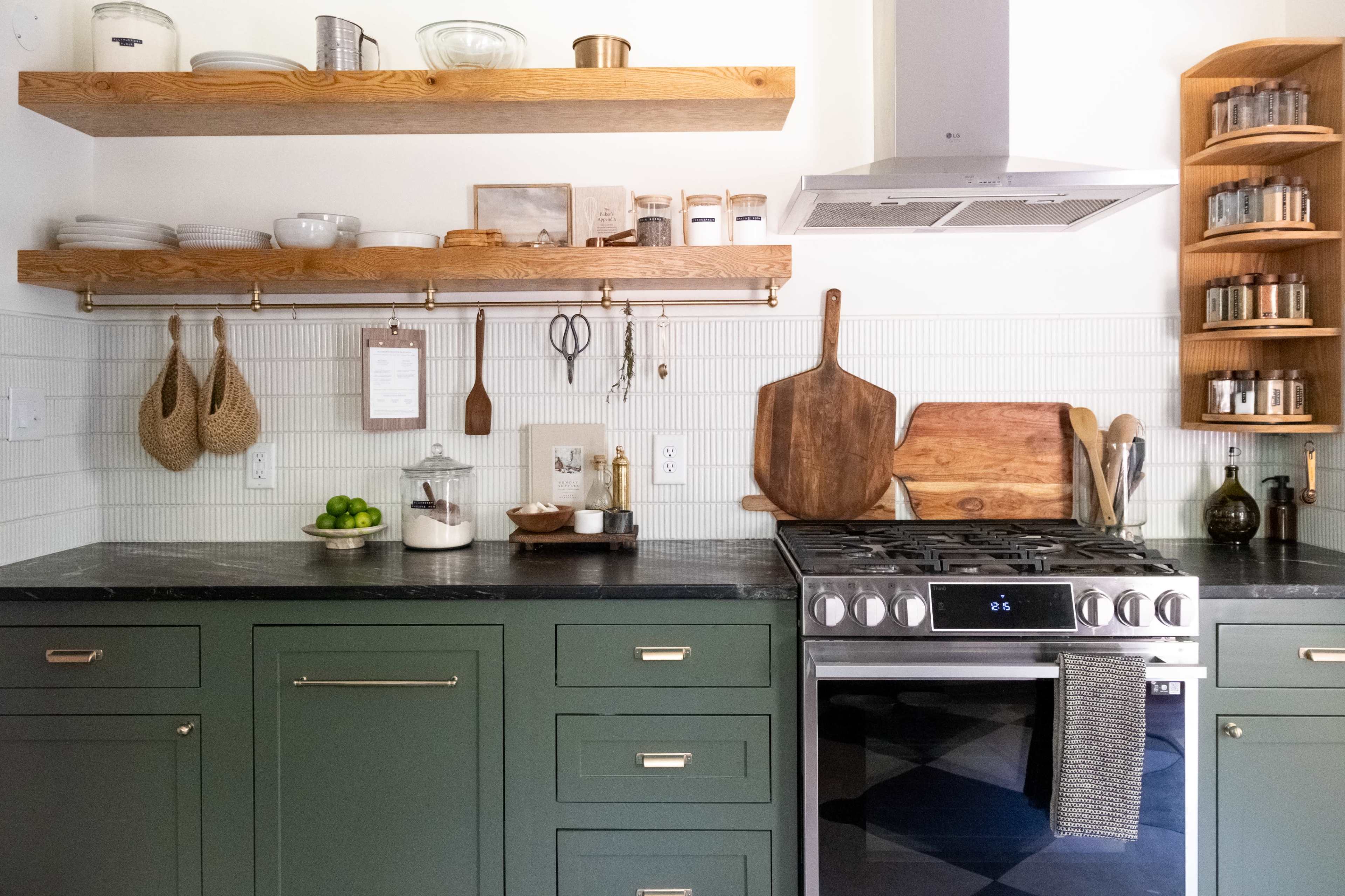 A modern kitchen features green cabinetry, wooden shelves with kitchen utensils, and a stainless steel stove against a white tiled backsplash.