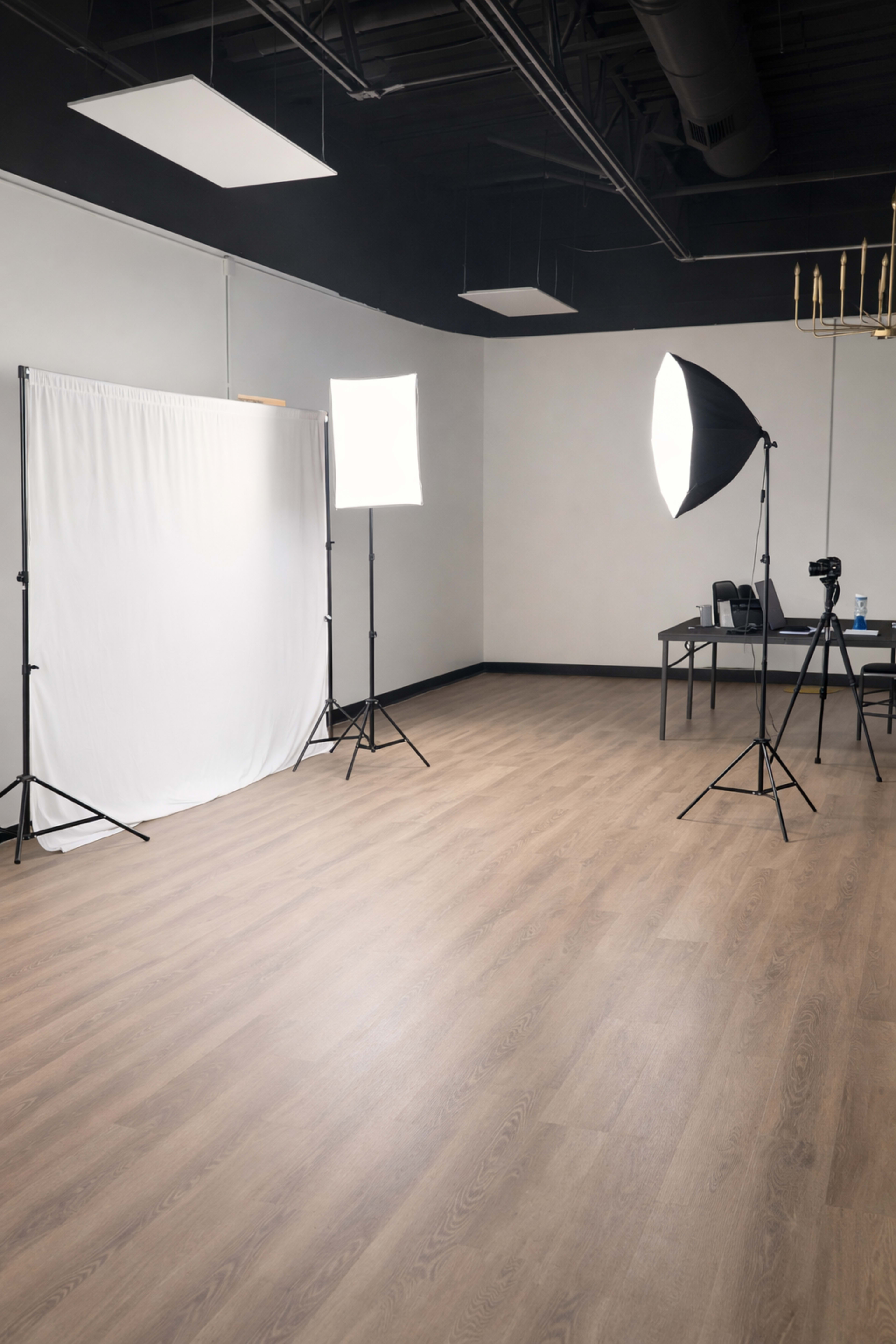 The image shows a photo studio with a white backdrop, two light sources, and a table with a camera set up in the corner.