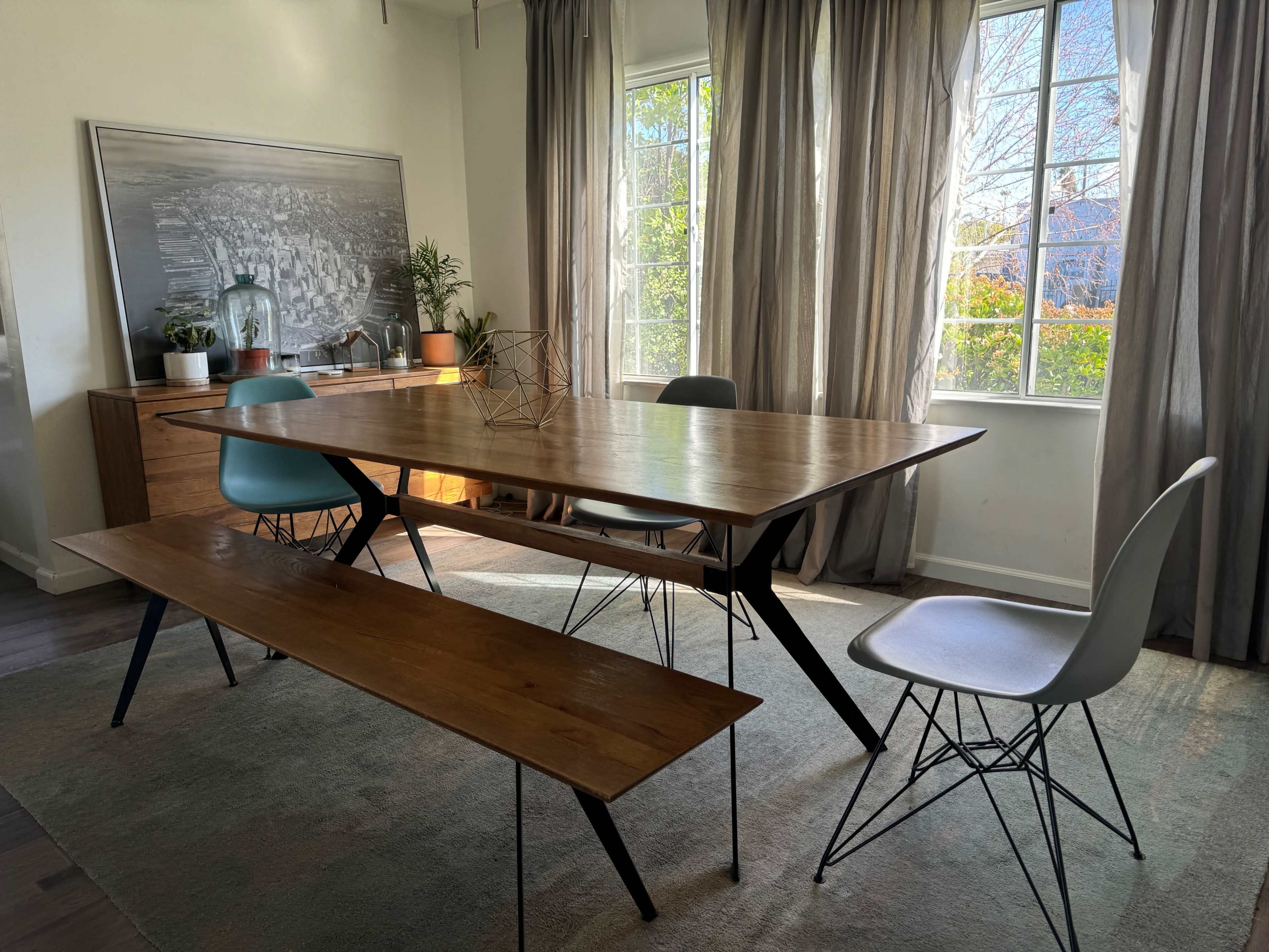 A wooden dining table with four chairs and a bench is positioned in a brightly lit room with large windows and decorative curtains.