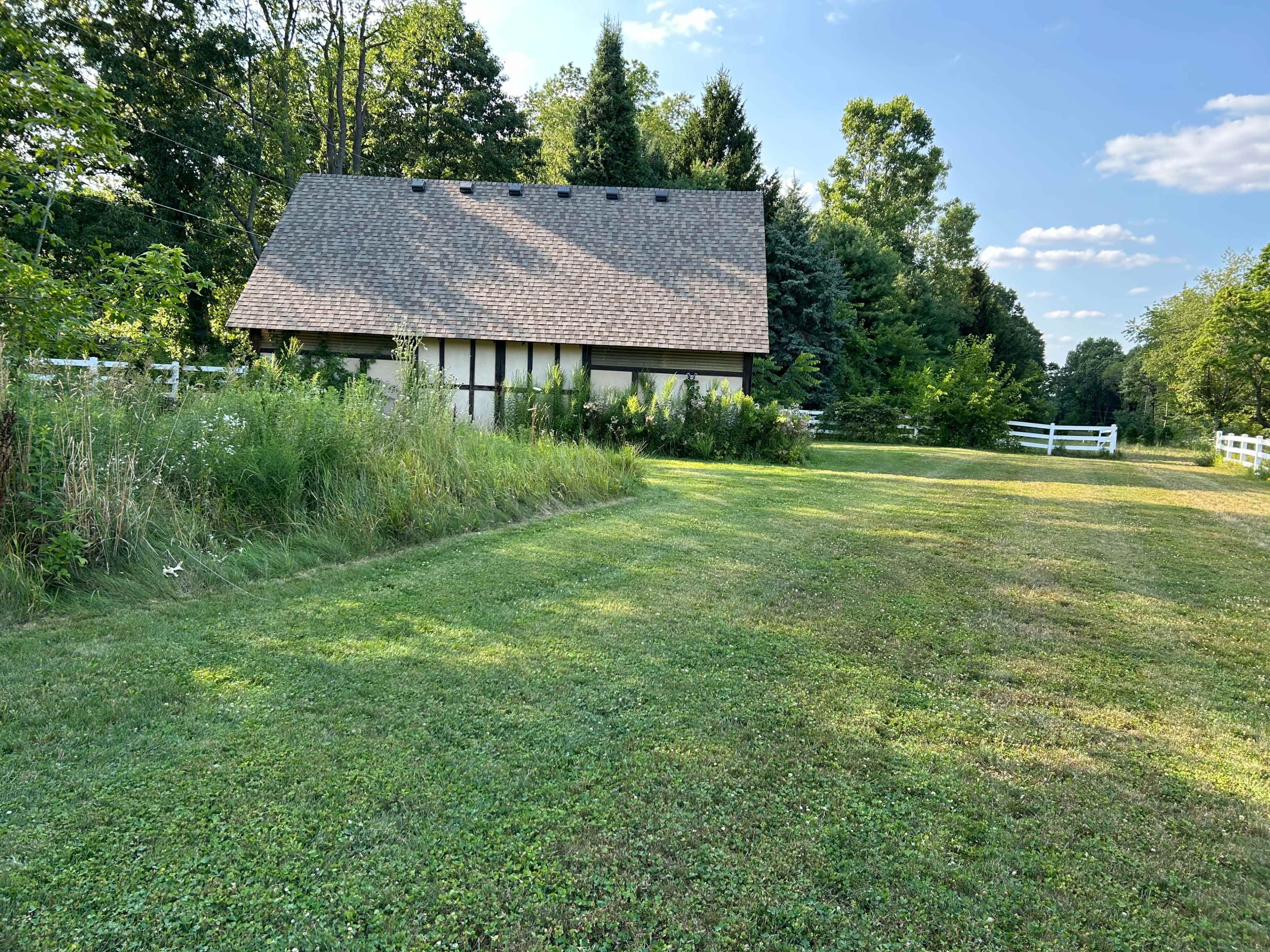 A rustic house with a sloped roof sits surrounded by greenery and a neatly mowed lawn, with a white fence in the background.