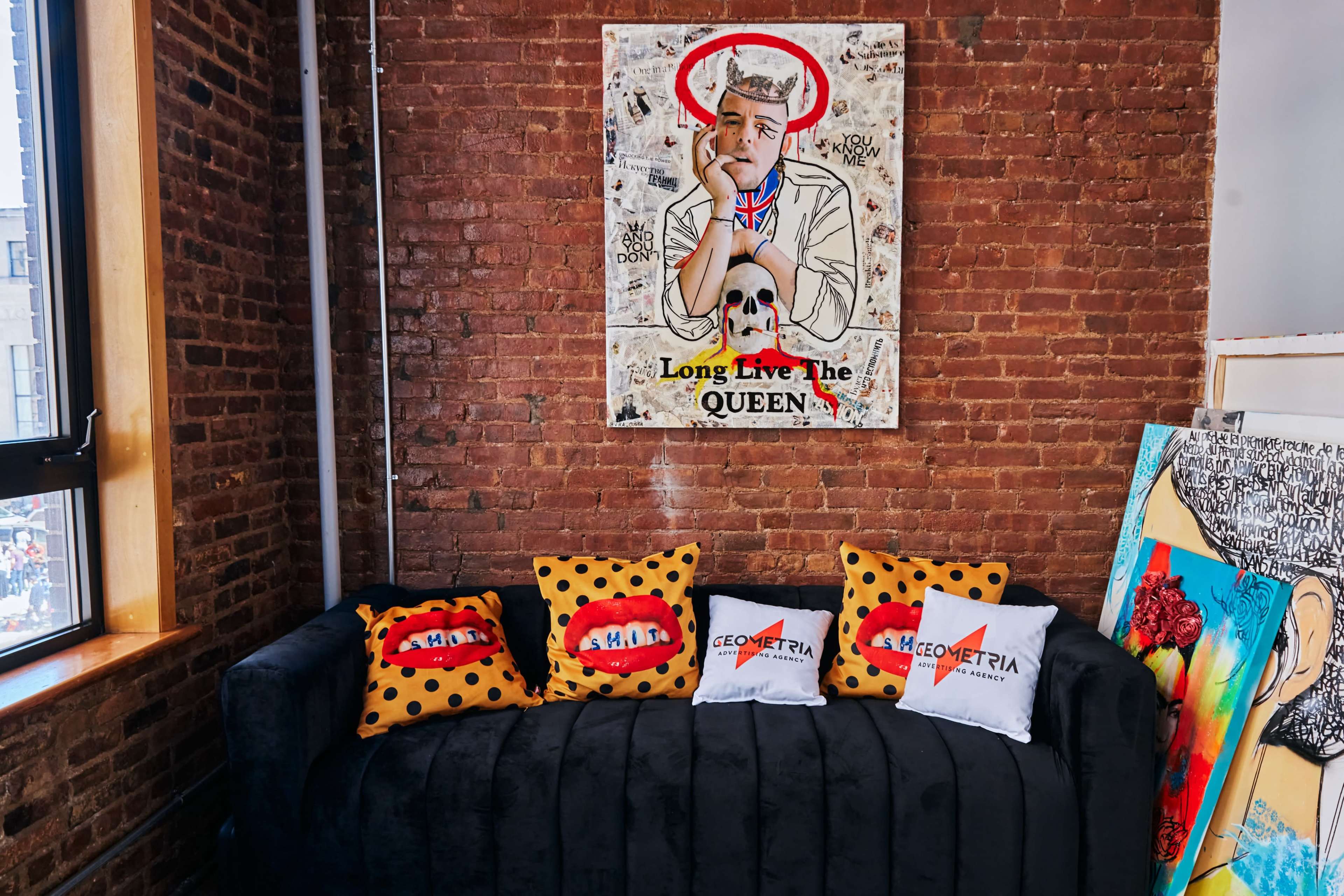 A black couch with patterned pillows, placed against a brick wall featuring a colorful art piece with the text "Long Live the QUEEN."