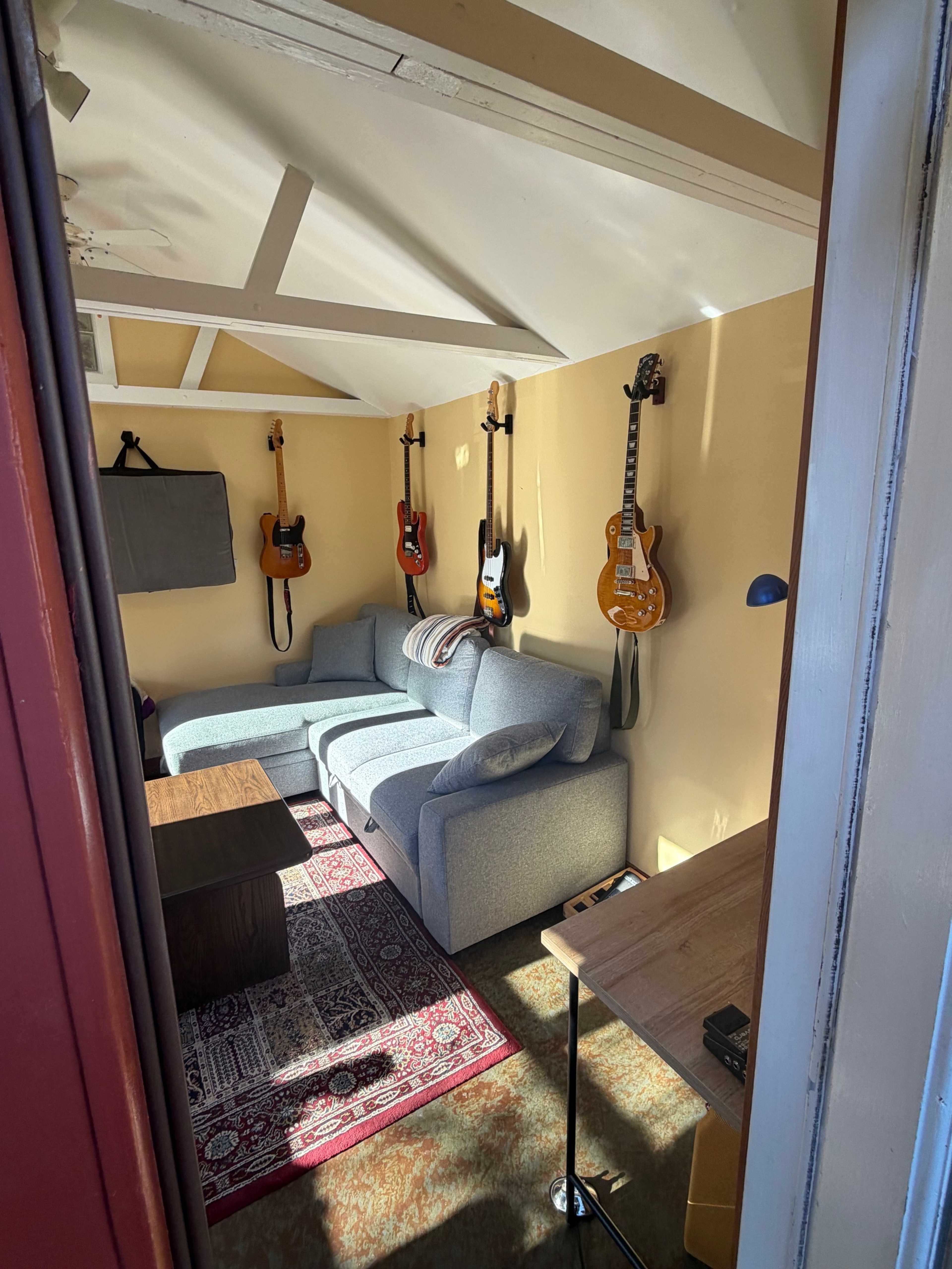 The image shows a small room with a gray sofa, a wooden table, and several guitars hanging on the walls.
