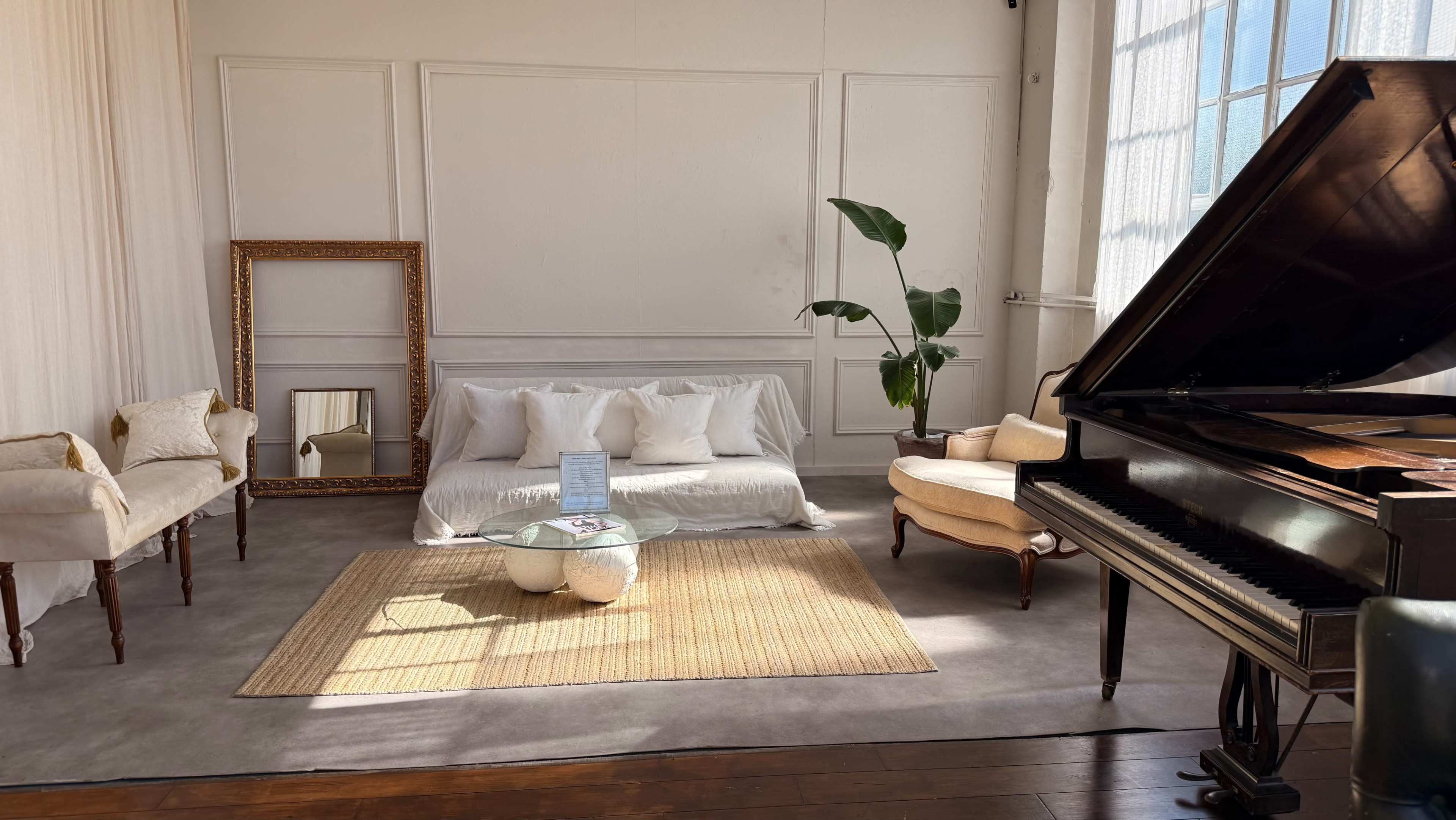 The scene features a bright room with a white sofa adorned with pillows, a vintage armchair, a grand piano, a glass coffee table, and a large mirror, all arranged over a neutral-colored rug.