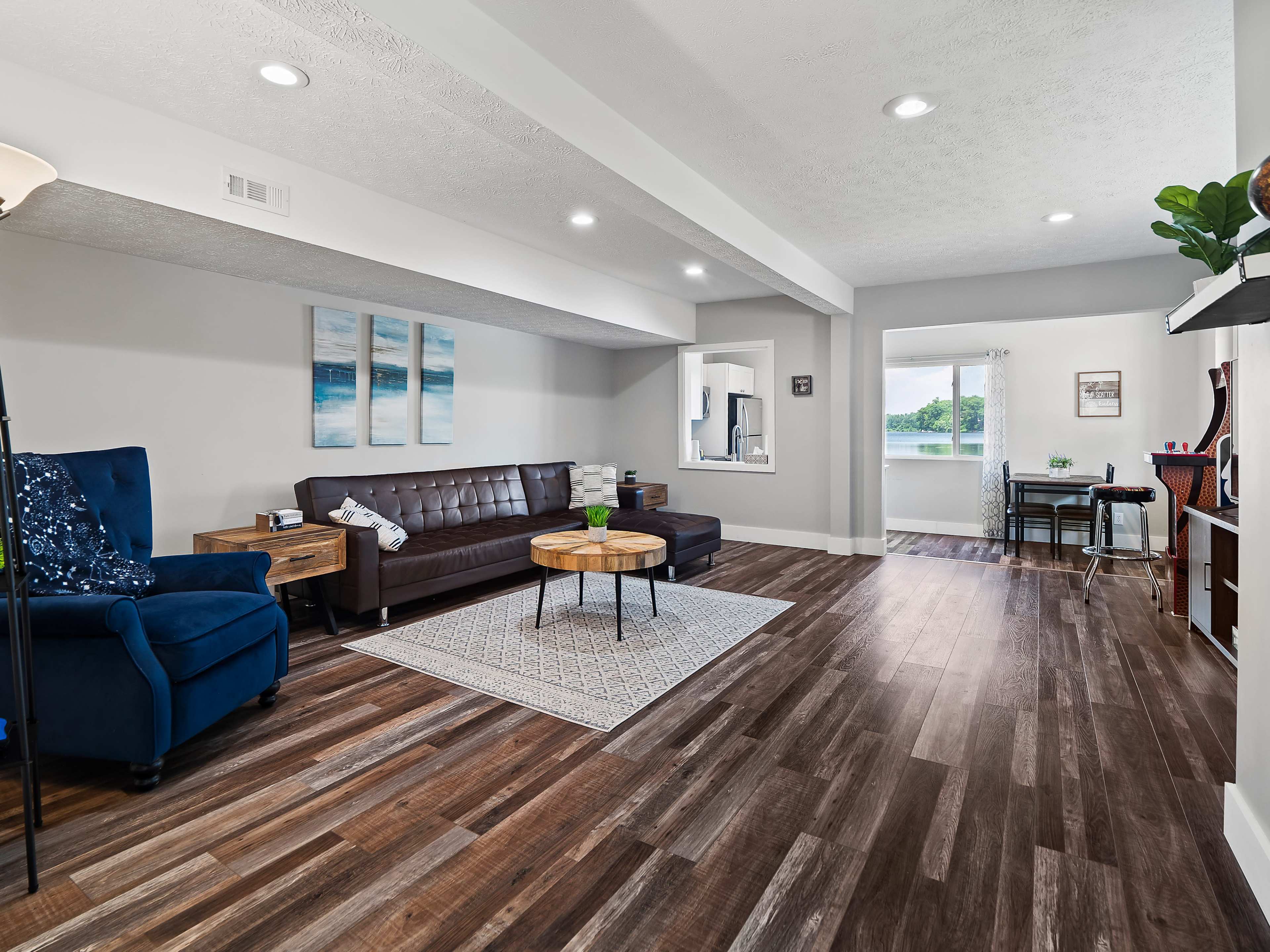 A spacious living area with brown hardwood floors, a dark brown leather sofa, a blue armchair, a round wooden coffee table, and a small desk by a window.